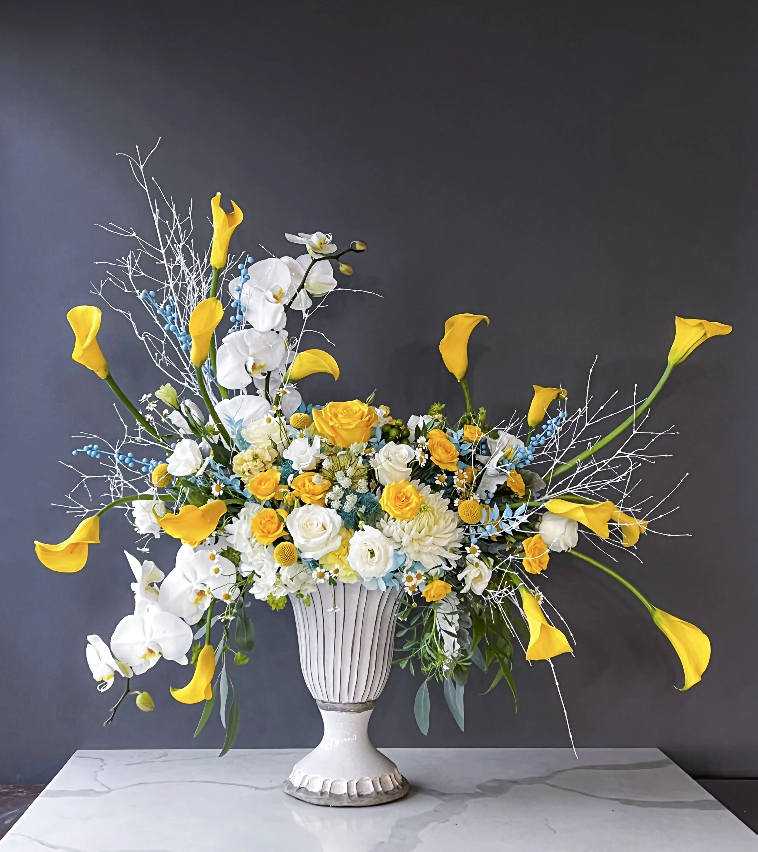 A large floral arrangement with yellow calla lilies, white orchids, yellow roses, and other flowers in a decorative white vase on a marble surface against a dark grey background.