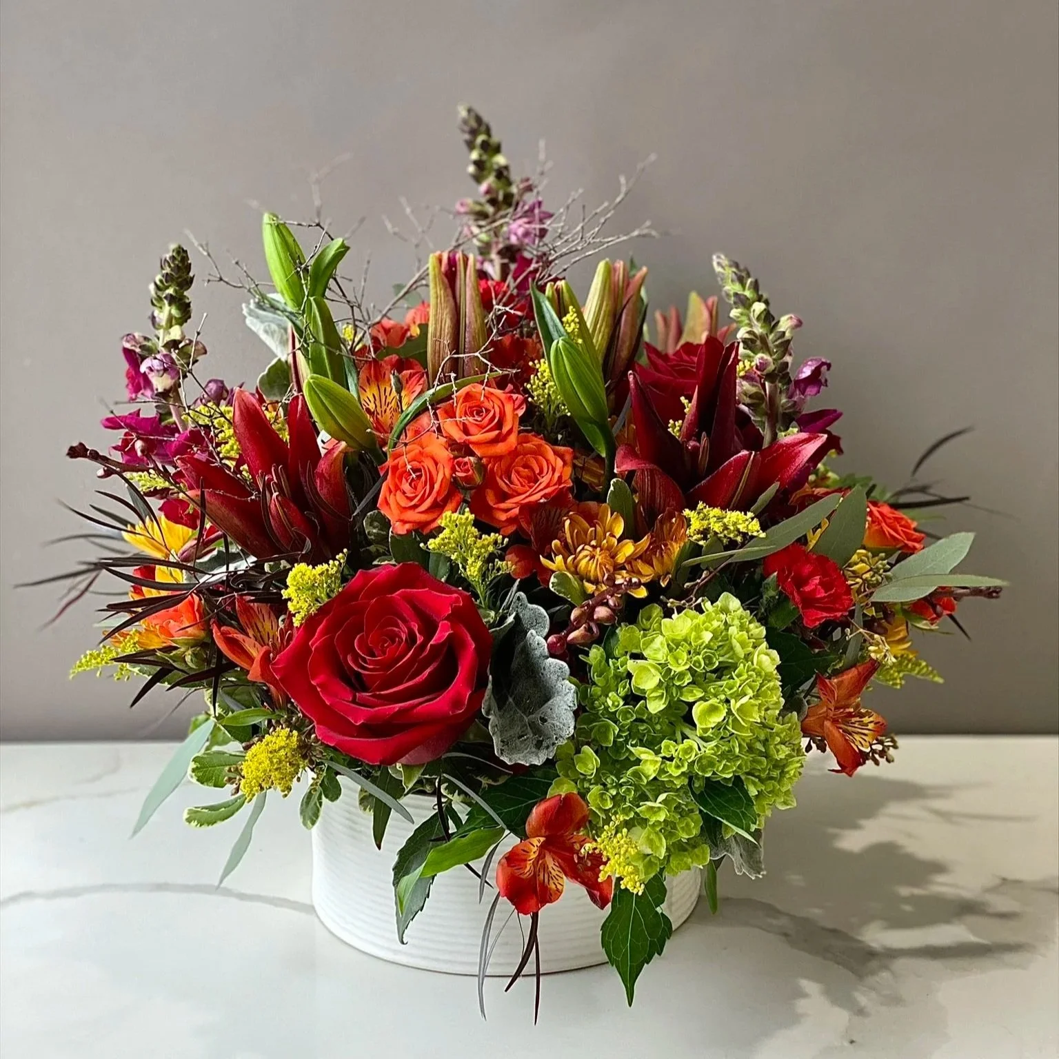 A colorful bouquet of flowers in a white vase, featuring red roses, orange roses, purple lilies, yellow chrysanthemums, green hydrangeas, and various green and purple foliage, placed on a white surface against a plain grey background.