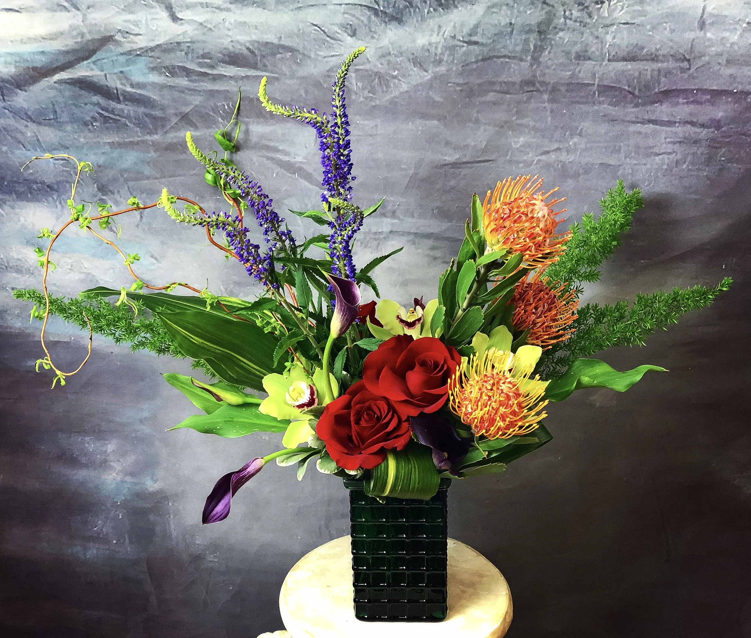 A colorful floral arrangement with purple, orange, red, yellow, and green flowers and foliage in a black geometric vase, placed on a gold-colored table against a dark gray textured background.