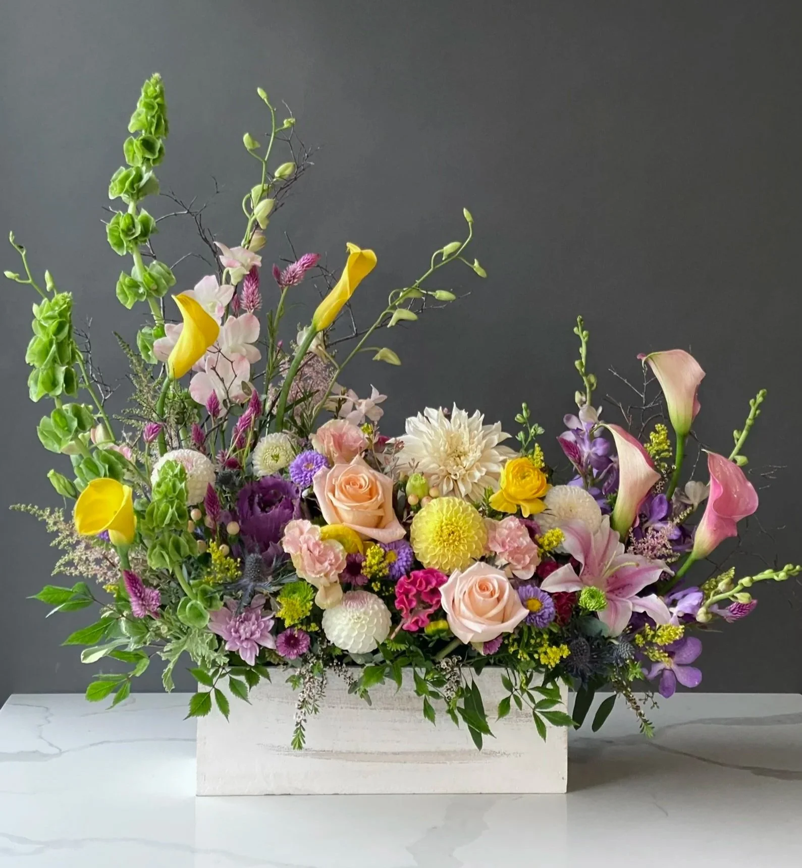 A floral arrangement with a variety of colorful flowers in a white container on a white surface against a dark background.