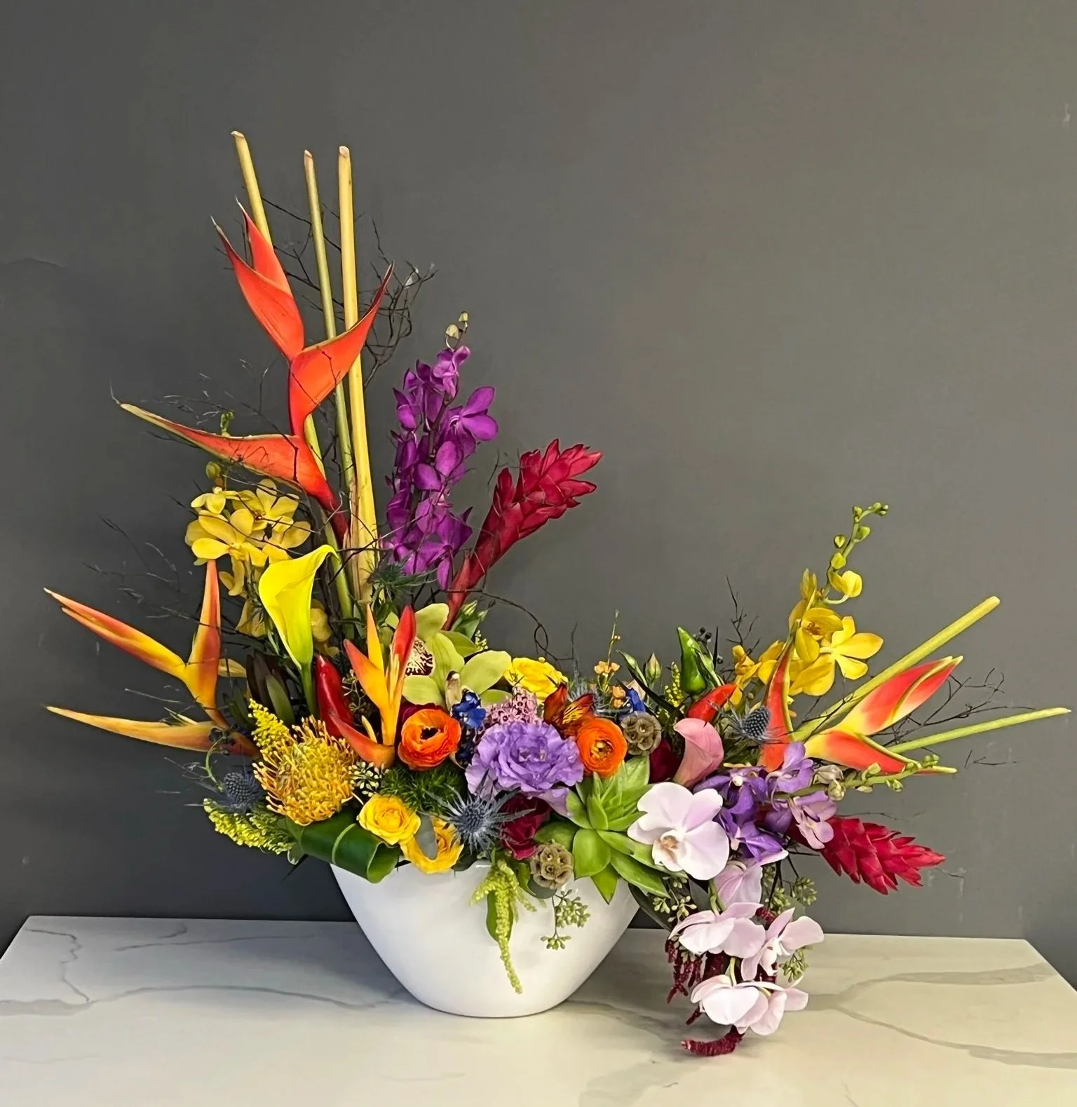 A vibrant mixed flower arrangement in a white ceramic vase featuring orange, yellow, purple, pink, and red flowers, arranged against a dark grey background.