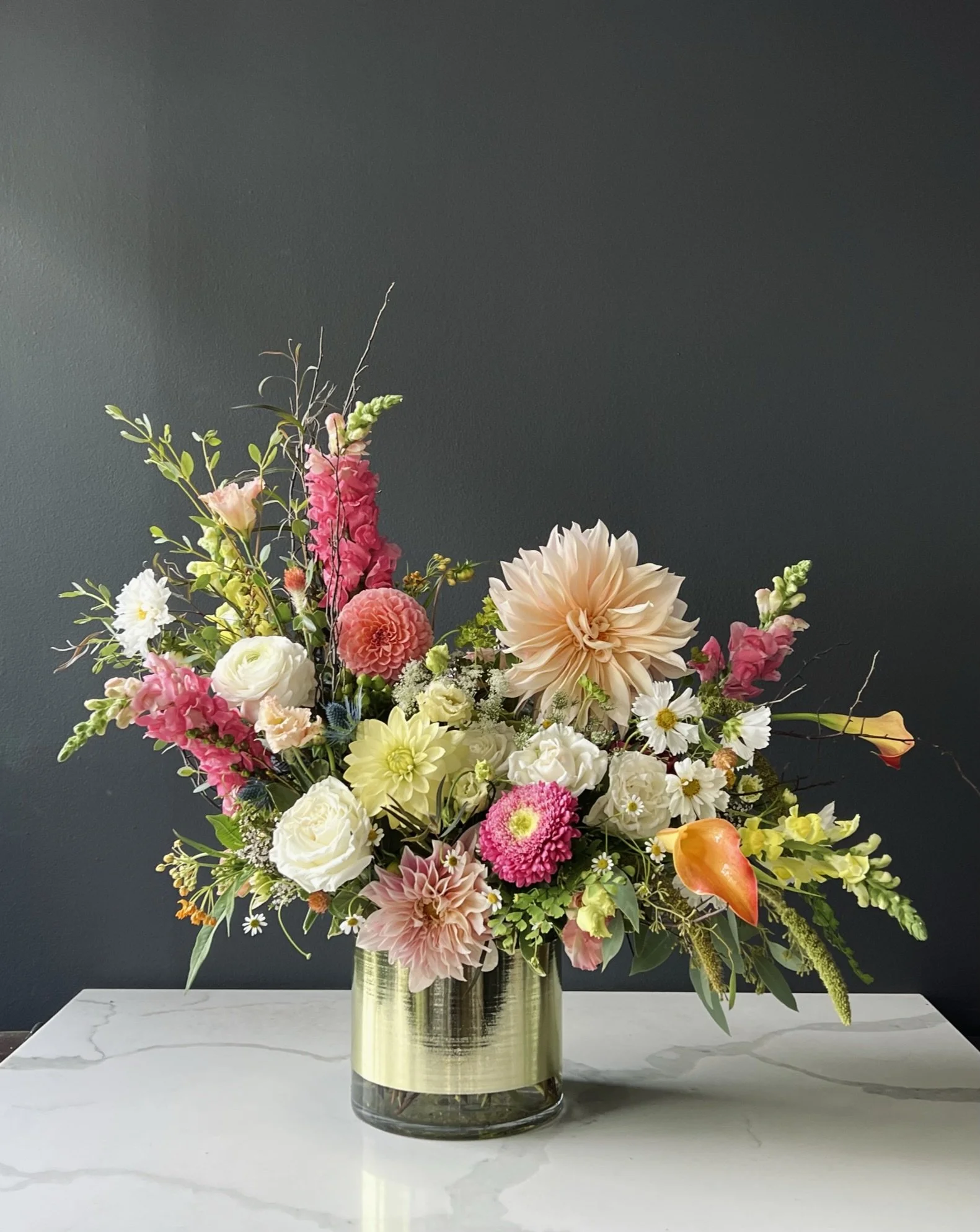 A large colorful flower bouquet in a light-colored cylindrical vase on a white marble surface against a dark background.