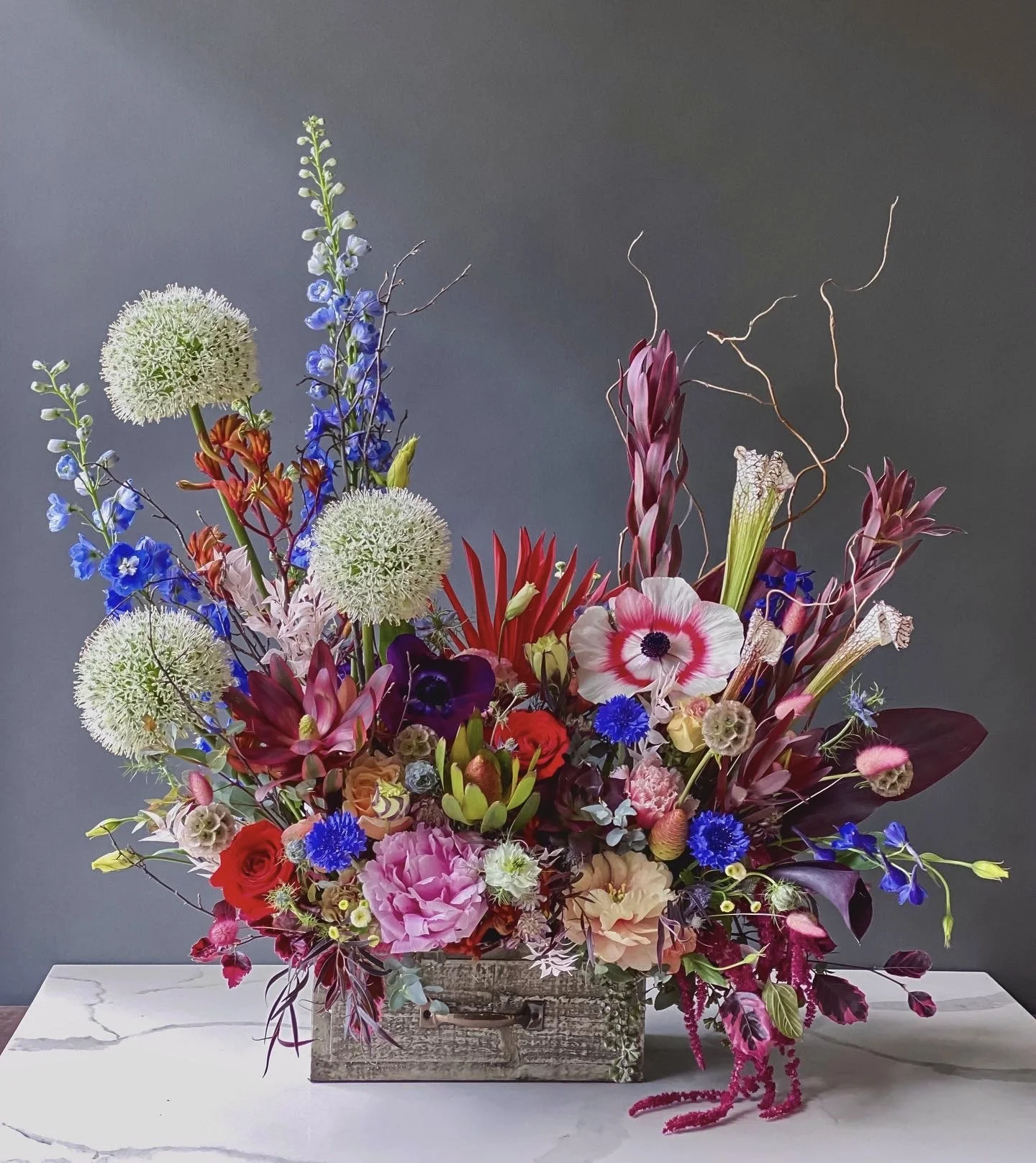 A vibrant bouquet of various colorful flowers arranged in a rustic wooden box, set against a dark gray background and placed on a white marble surface.