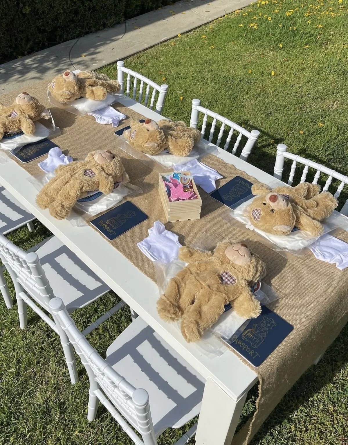 A long white rectangular table set outdoors with six teddy bears placed on top, each lying on a pillow, surrounded by cups, napkins, and a small stack of pink cutouts in the center. The table has white chairs around it, and the setting is on a grassy lawn with a concrete path nearby.