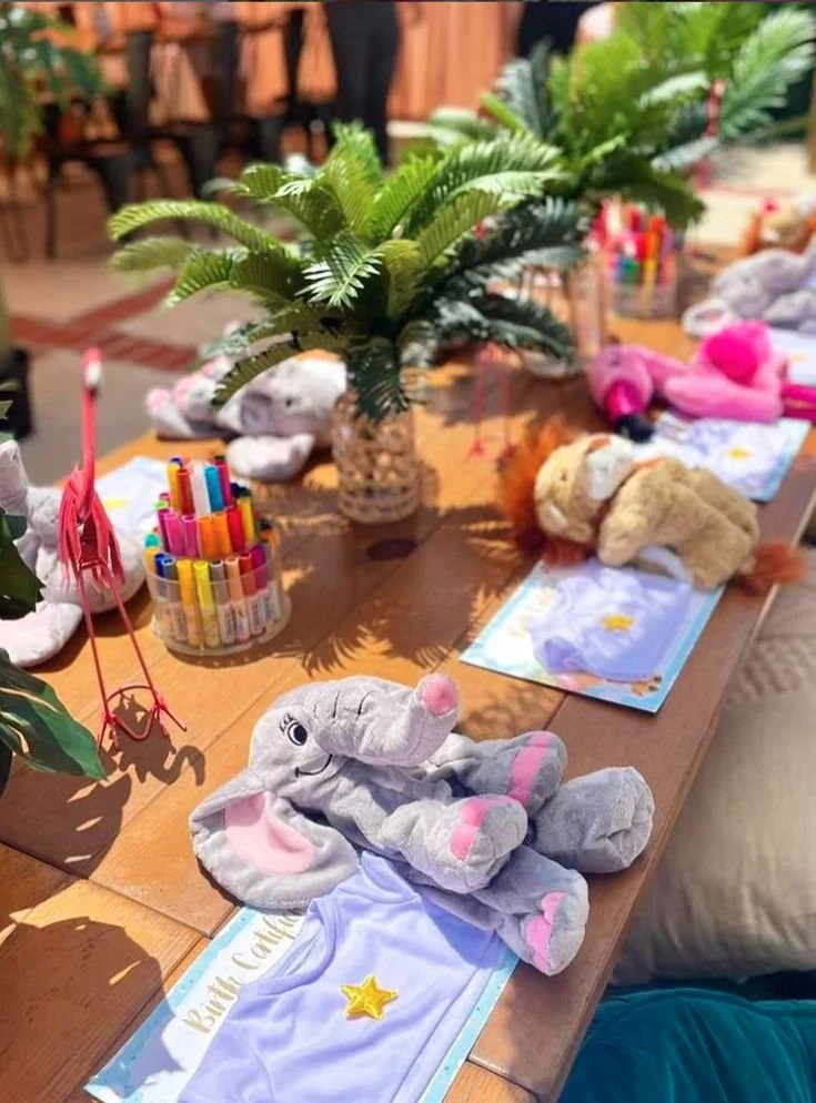 A table decorated with plush animal toys, coloring supplies, and paper placemats in a party setting. Central is a potted plant, with plush elephants and lions on the table.