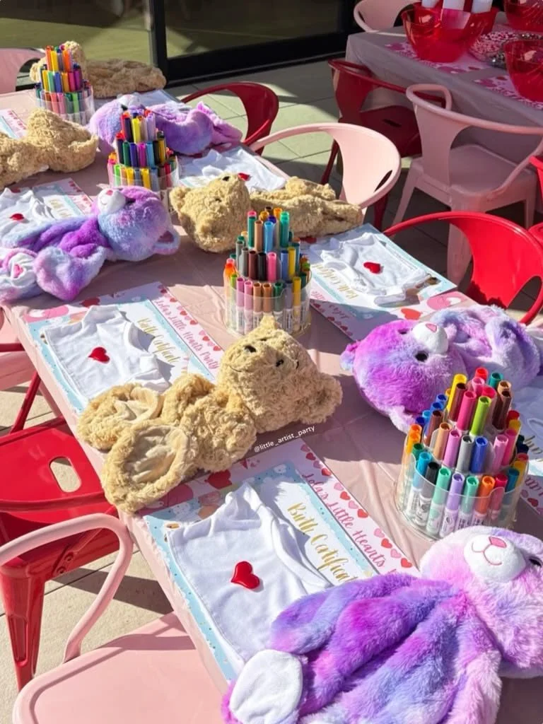 Decorated table with plush bear and purple bunny stuffed animals, colorful markers in containers, and pink and red tableware, set for a children's party.