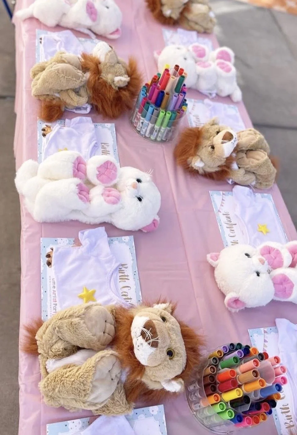 Table with stuffed animal plush toys, including lions and rabbits, arranged for a craft activity with colorful markers in the center on a pink tablecloth.
