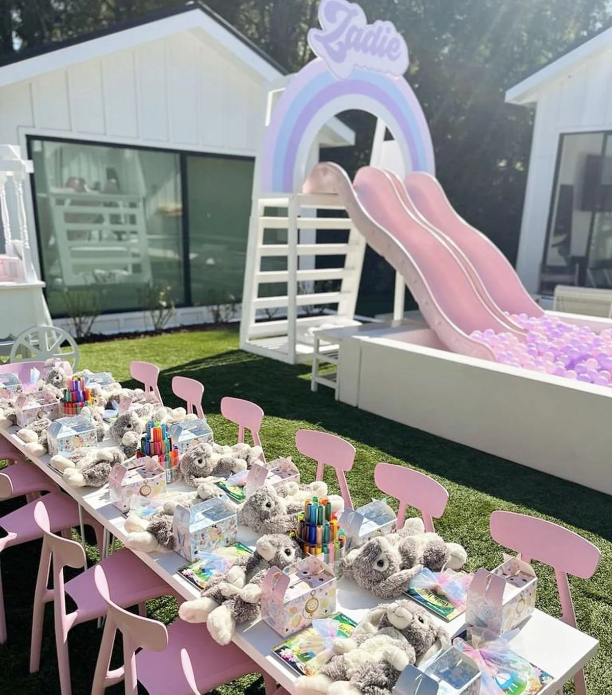 A children's birthday party setup outdoors with a pink and purple rainbow-themed slide, a ball pit, and a table filled with teddy bears, coloring supplies, and party favors.