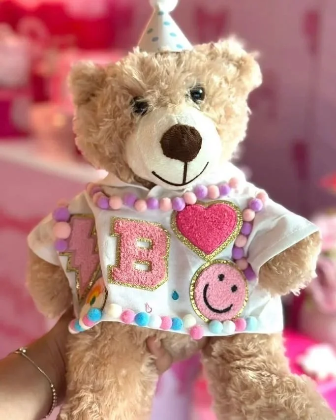 A teddy bear wearing a party hat and decorated with colorful patches and beads, including a pink heart, a smiling face, and the letter E on a pink patch, held in a person's hand against a pink background.