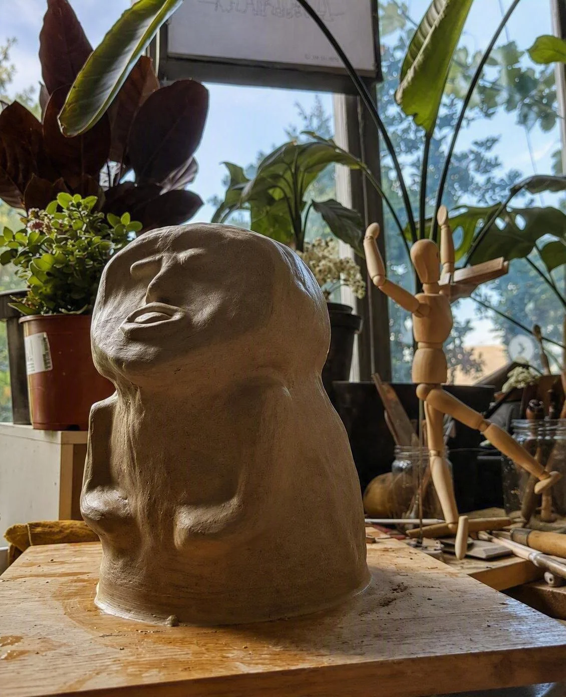 A ceramic sculpture of a human head on a wooden table, surrounded by plants, a wooden mannequin, and art supplies in a bright room with large windows.