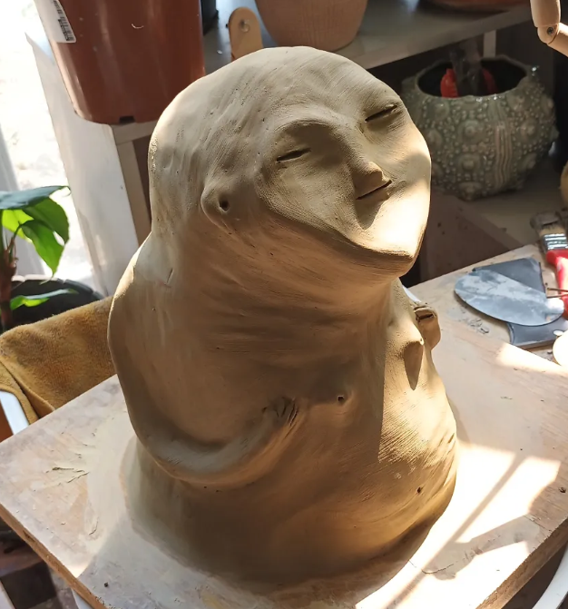 A clay sculpture of a smiling, relaxed face with closed eyes, sitting on a worktable in a studio.