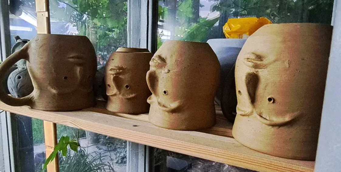 Four clay mugs shaped like human torsos with facial features, arranged on a wooden shelf in front of a garden window.