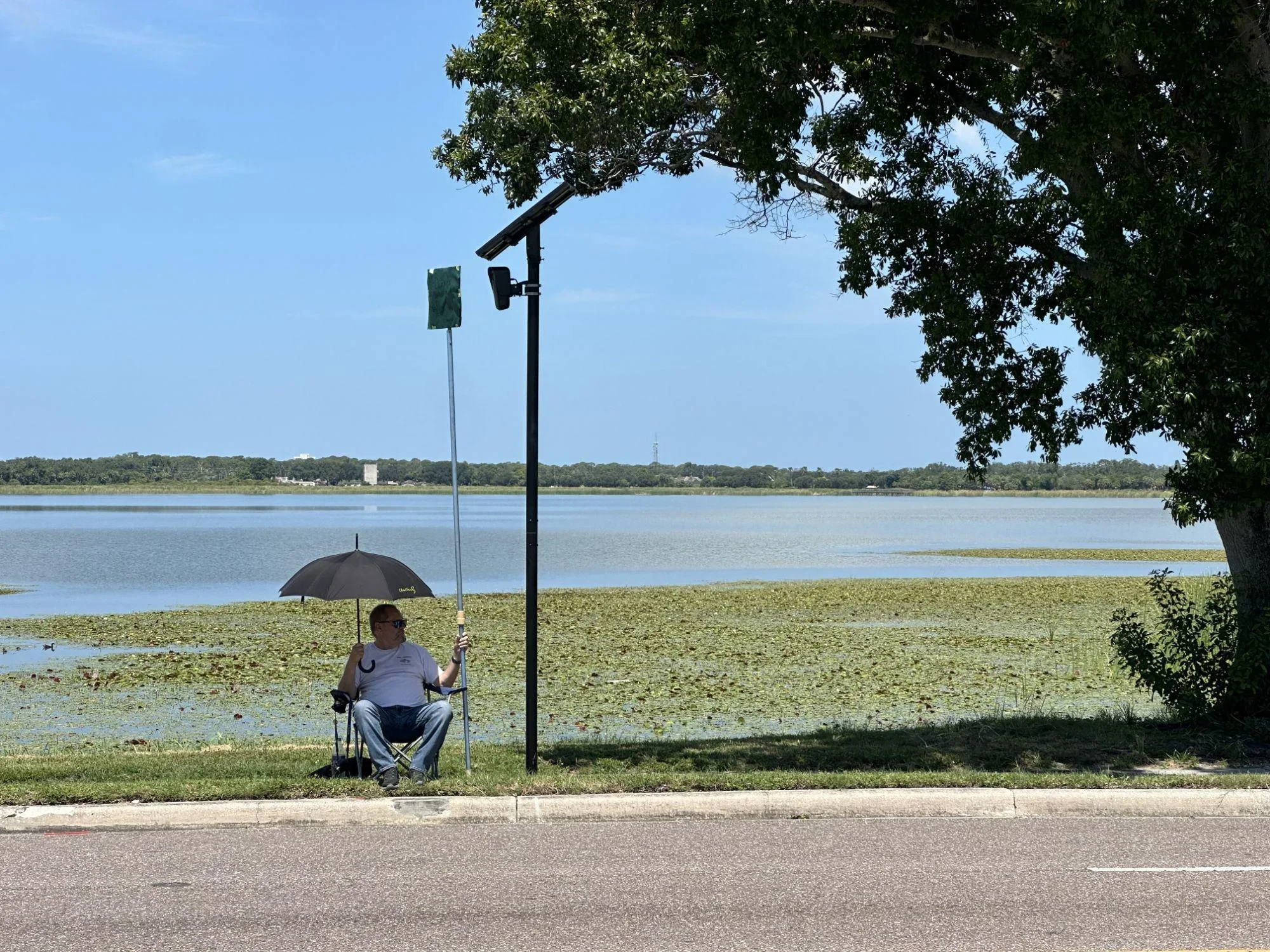 A man sitting in a lawn chair holding a sign in front of an ALPR Flock camera to block it from surveilling passing traffic.