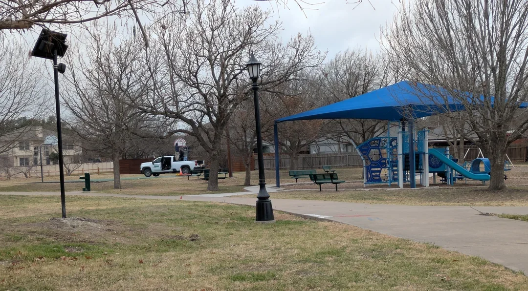 Public park with a Flock Safety ALPR camera facing a playground under a blue canopy, benches, a lamp post, leafless trees, and a truck parked in the background.