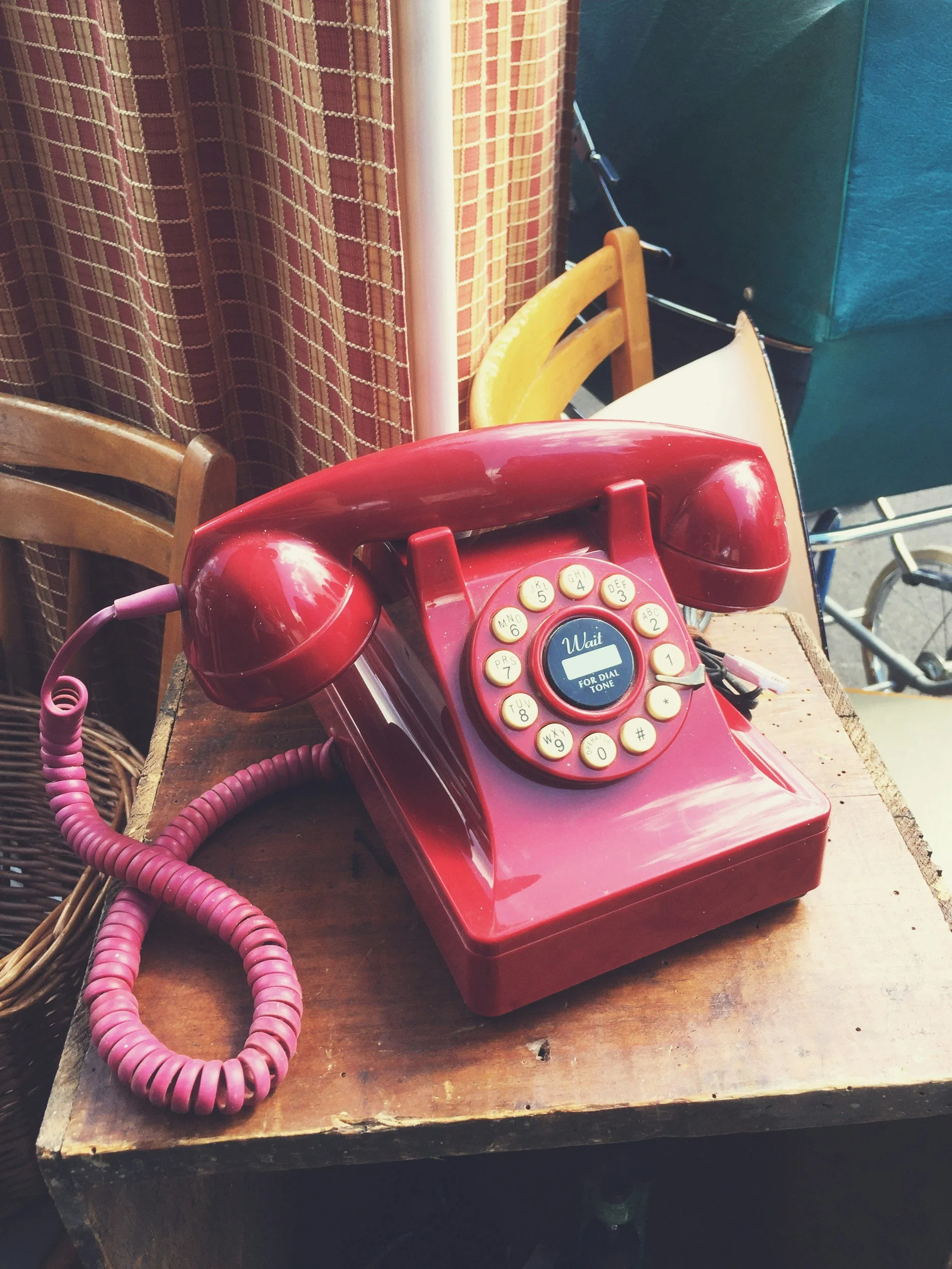 A red vintage rotary phone with a coiled pink cord sitting on a wooden surface. Behind it, there are various chairs and a patterned curtain.