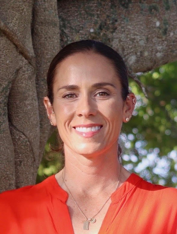 A woman with brown hair and a bright smile standing outdoors next to a tree with green leaves in the background, wearing a red top and a silver necklace.