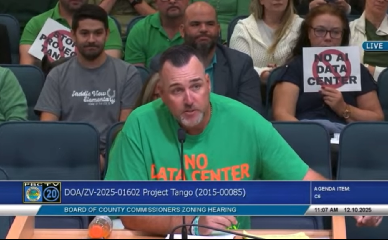 A man in a green shirt with the words 'NO LATE CENTER' is speaking into a microphone at a public zoning hearing, with several people seated behind him, some holding signs that say 'NO DATA CENTER'.