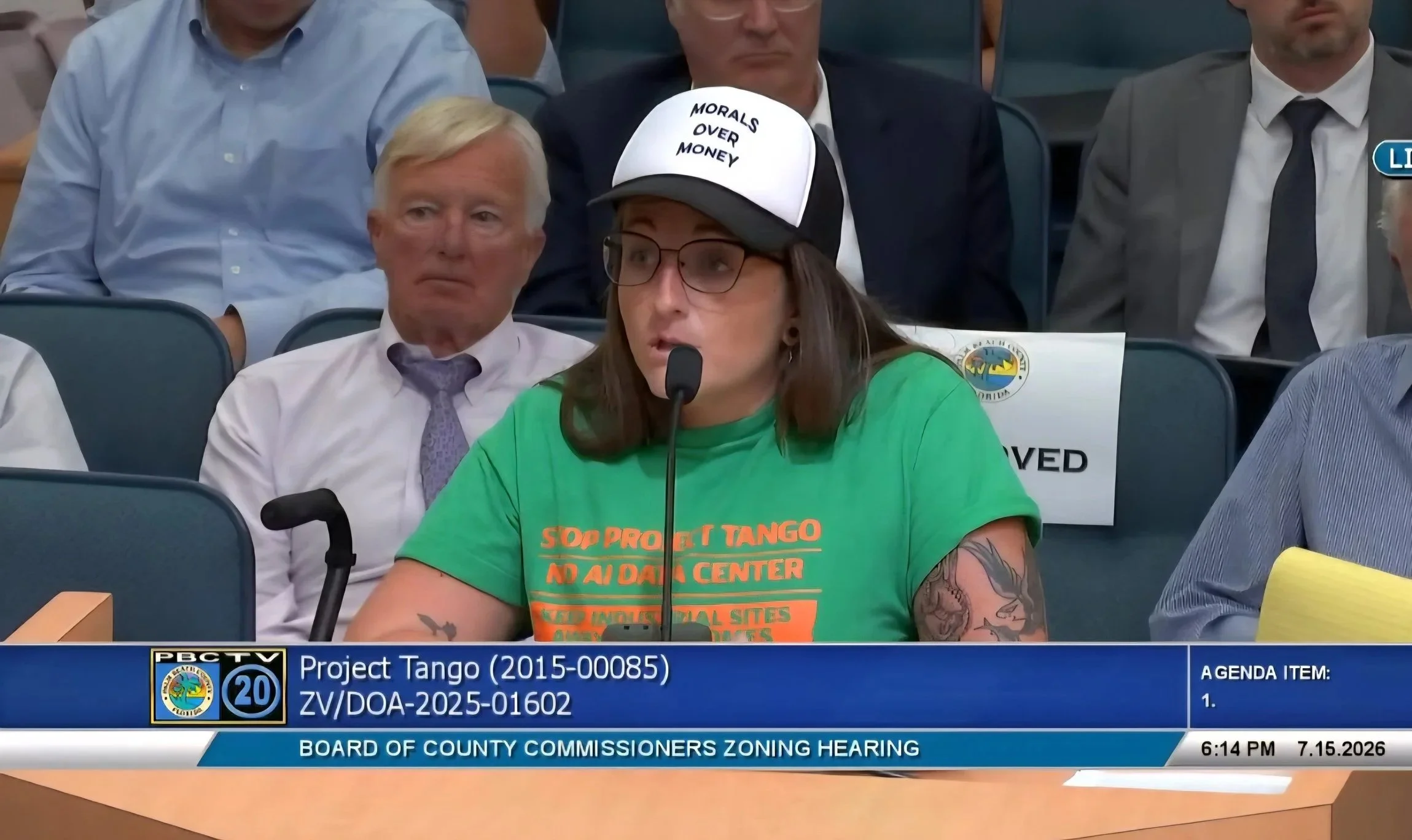 A woman speaking into a microphone at a county zoning hearing, wearing a cap that reads 'Morals Over Money' and a green T-shirt supporting Project Tango, with people seated behind her in a formal setting.