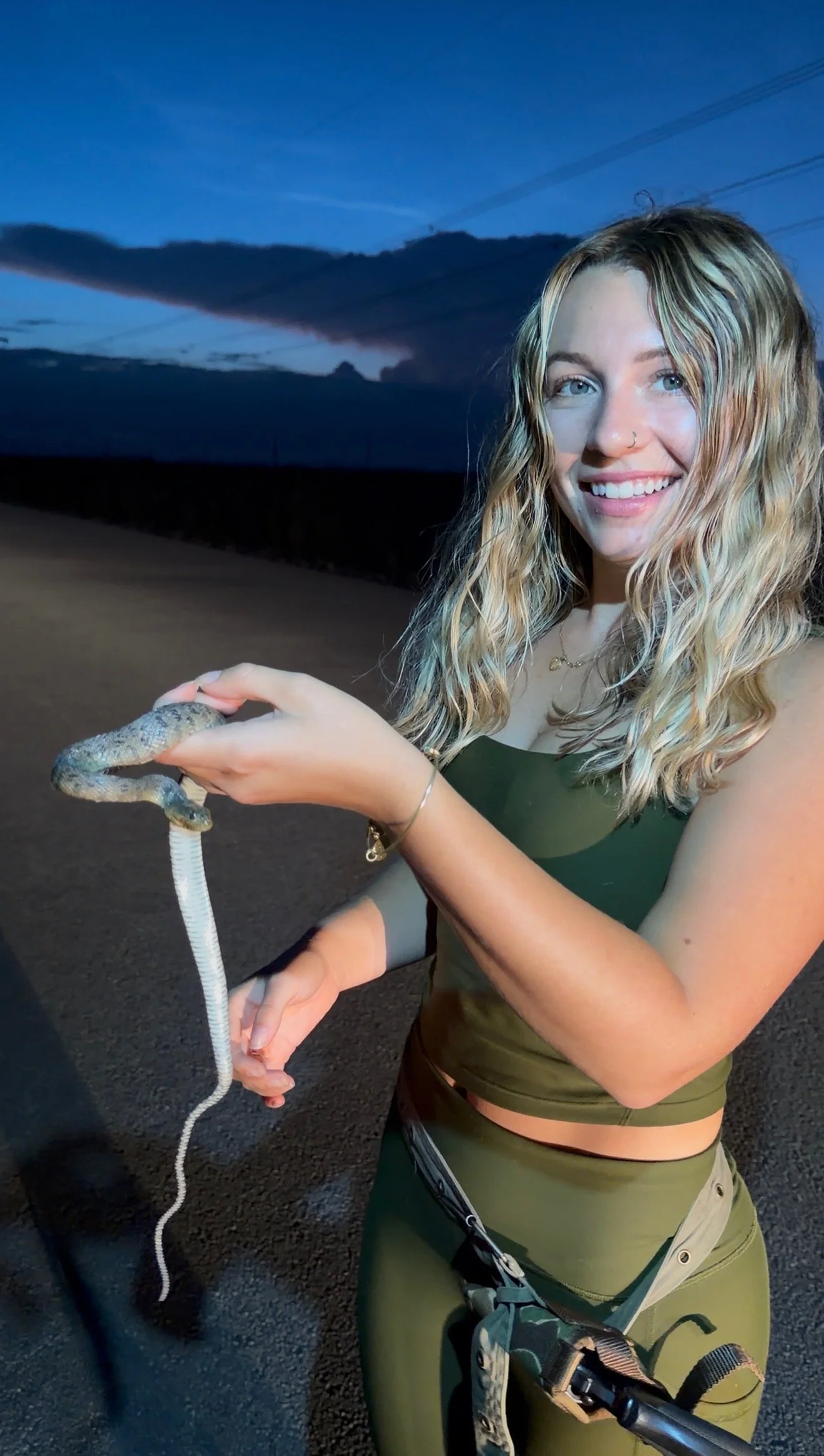A young woman with blonde wavy hair and wearing green athletic gear, smiling while holding a small snake with a pale body and darker head, outdoors during dusk with clouds in the sky.