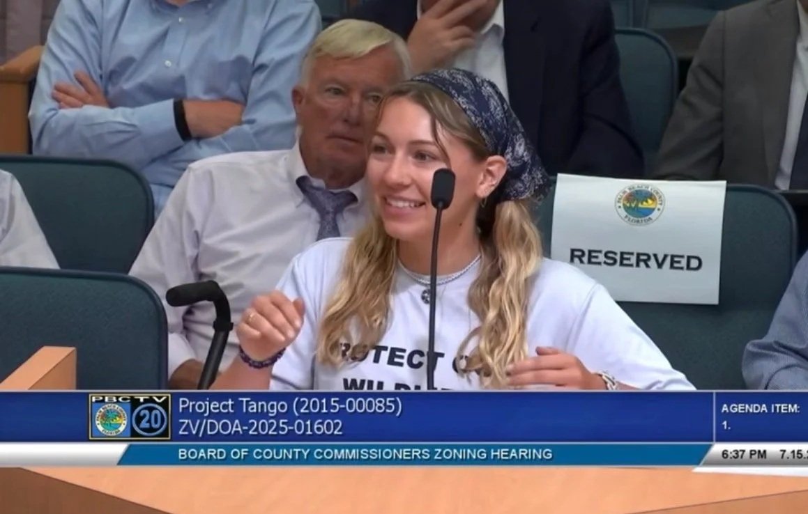 A young woman with blonde hair, wearing a bandana, speaking into a microphone at a county zoning hearing, smiling. She is surrounded by seated individuals, some taking notes, with a reserved sign visible on the seat behind her.