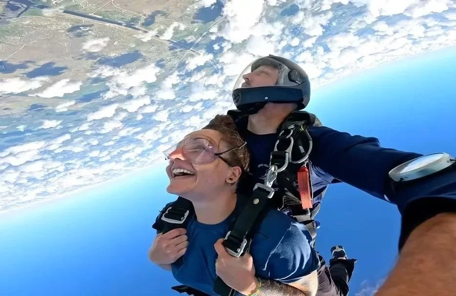 Two people skydiving, one with a big smile and the other wearing a helmet, free-falling above the clouds and land.