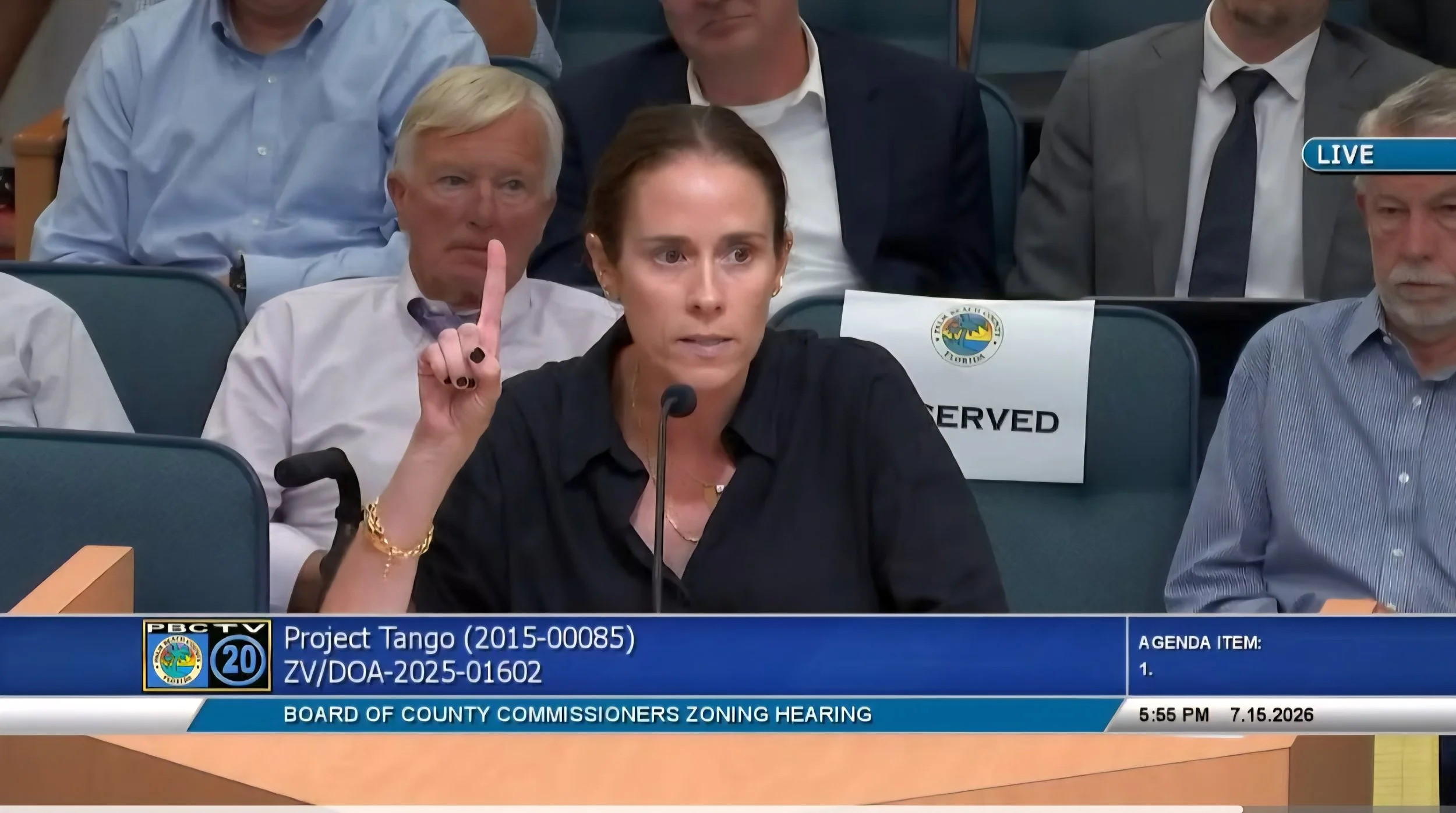 A woman speaking at a public hearing for the Board of County Commissioners, with several seated people behind her, including a man with white hair and a woman with glasses, in a formal setting with official signage and a timer displayed.