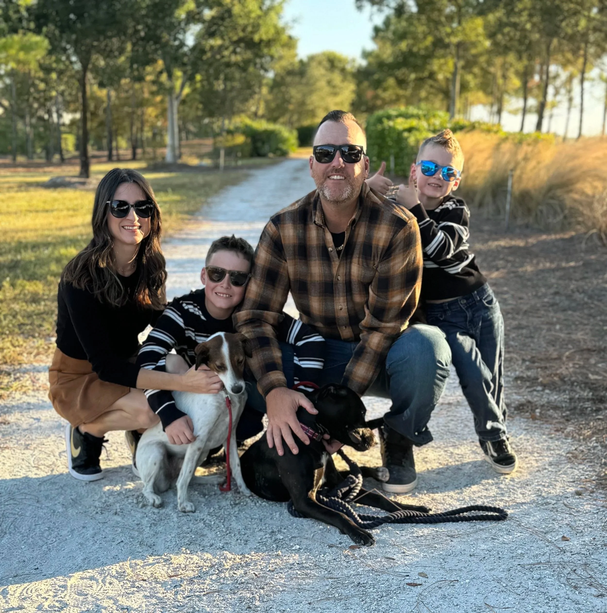 A group of five people, including adults and children, posing outdoors with a dog during what appears to be late afternoon or early evening. They are smiling and wearing sunglasses, with a background of green trees and a dirt path.