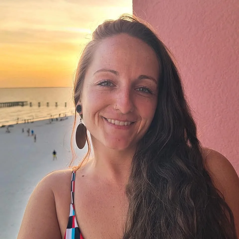 A woman smiling at sunset on a beach, wearing earrings and a colorful striped swimsuit, with waves and a pier in the background.