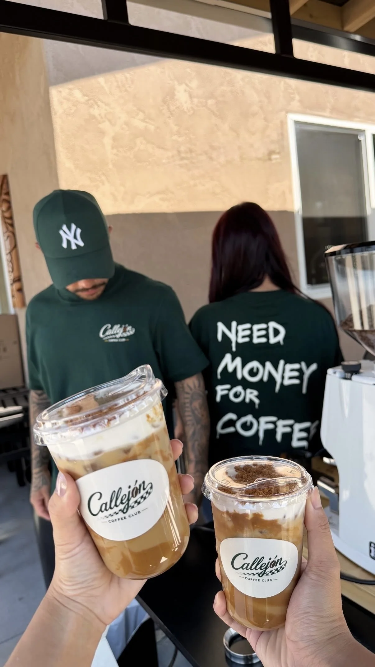 Two people holding coffee drinks with 'Callejón Coffee Club' labels at a coffee shop counter. A barista wearing a 'Need Money for Coffee' shirt stands behind the counter. The drinks have cold foam and toppings.