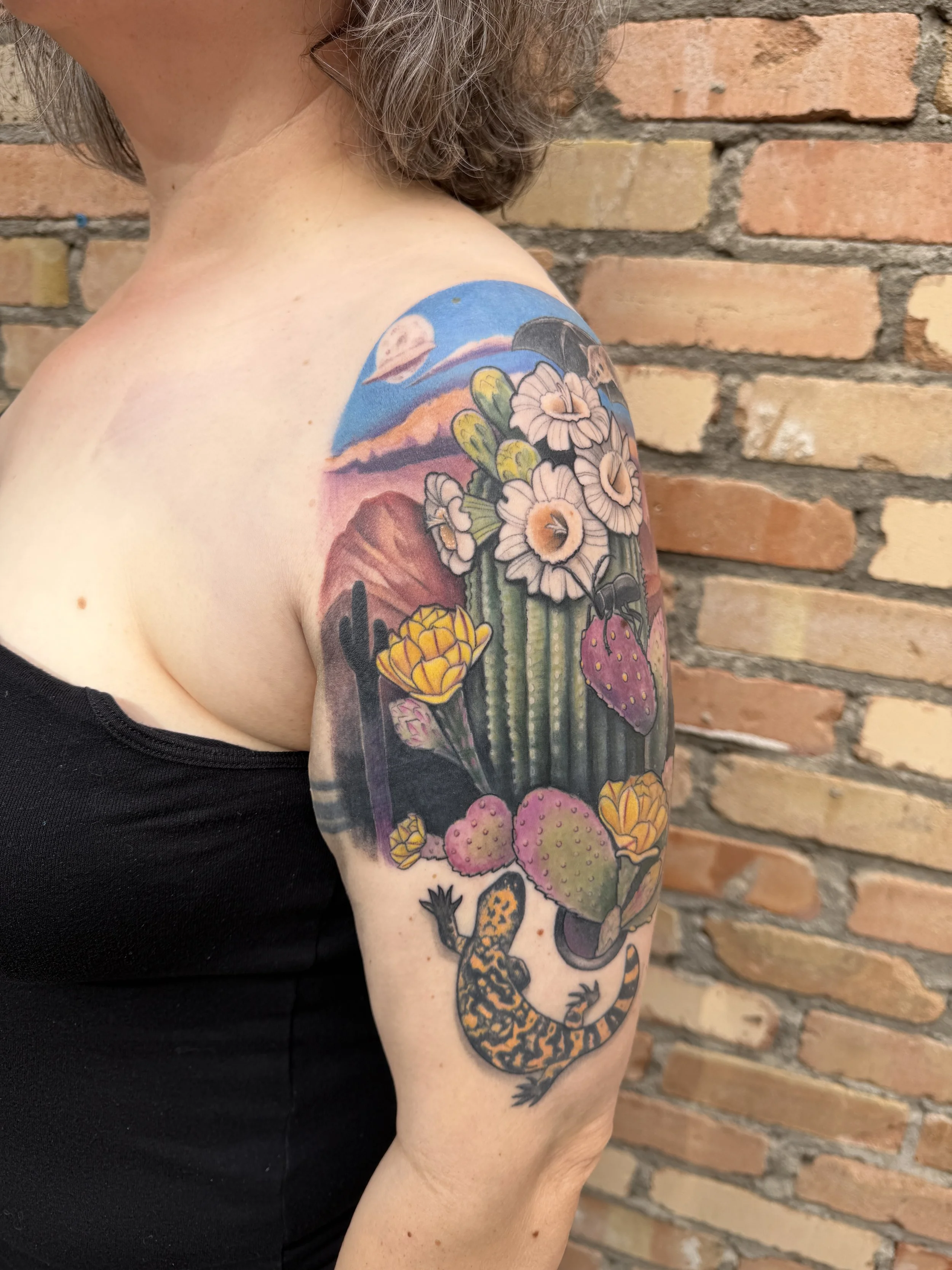 Close-up of a person's upper arm with a colorful tattoo of desert plants and cacti, a sunset or sunrise sky, and a moon, with a brick wall in the background.