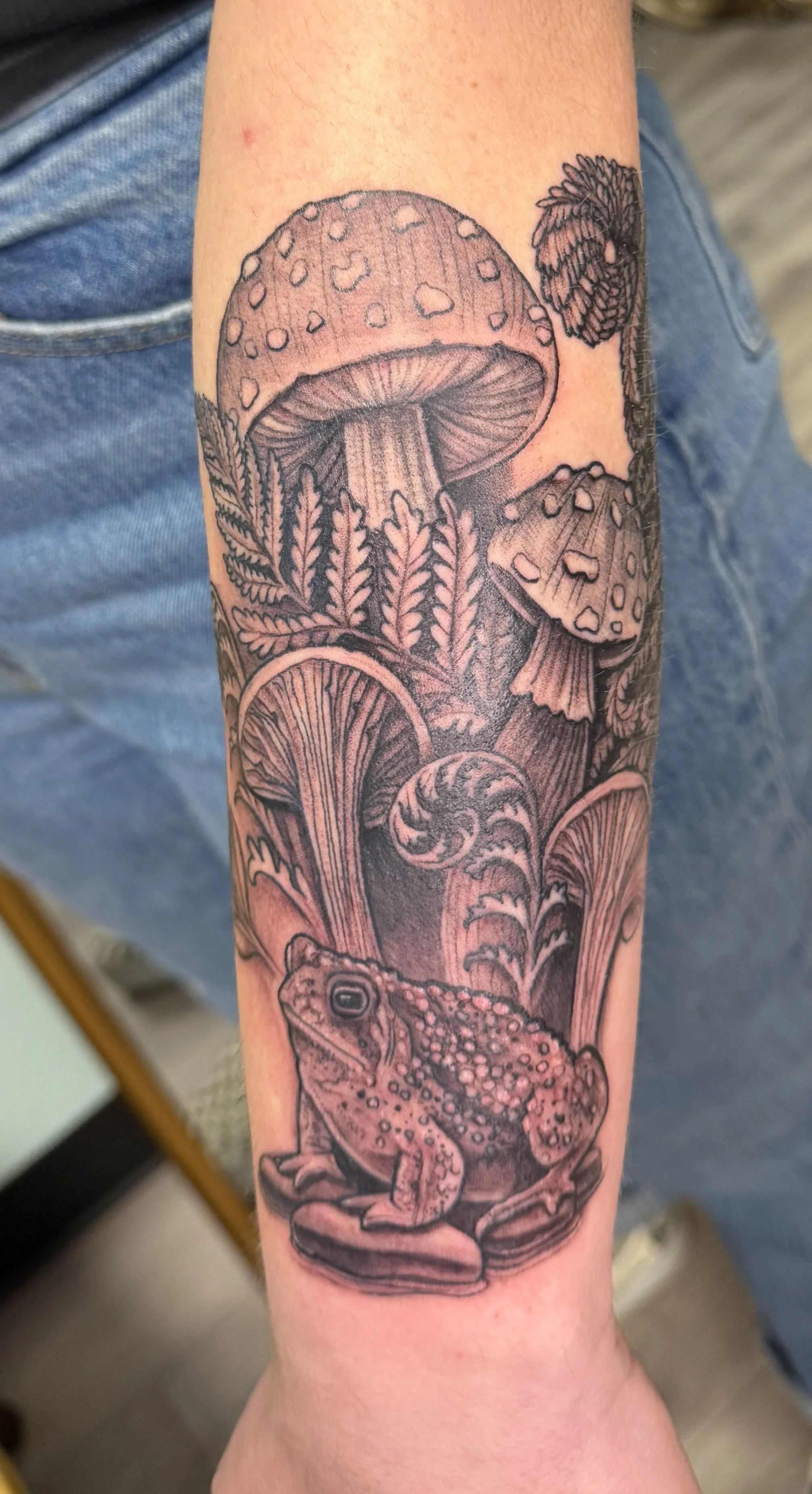 Black ink tattoo of mushrooms, fern leaves, and a frog on a person's forearm.