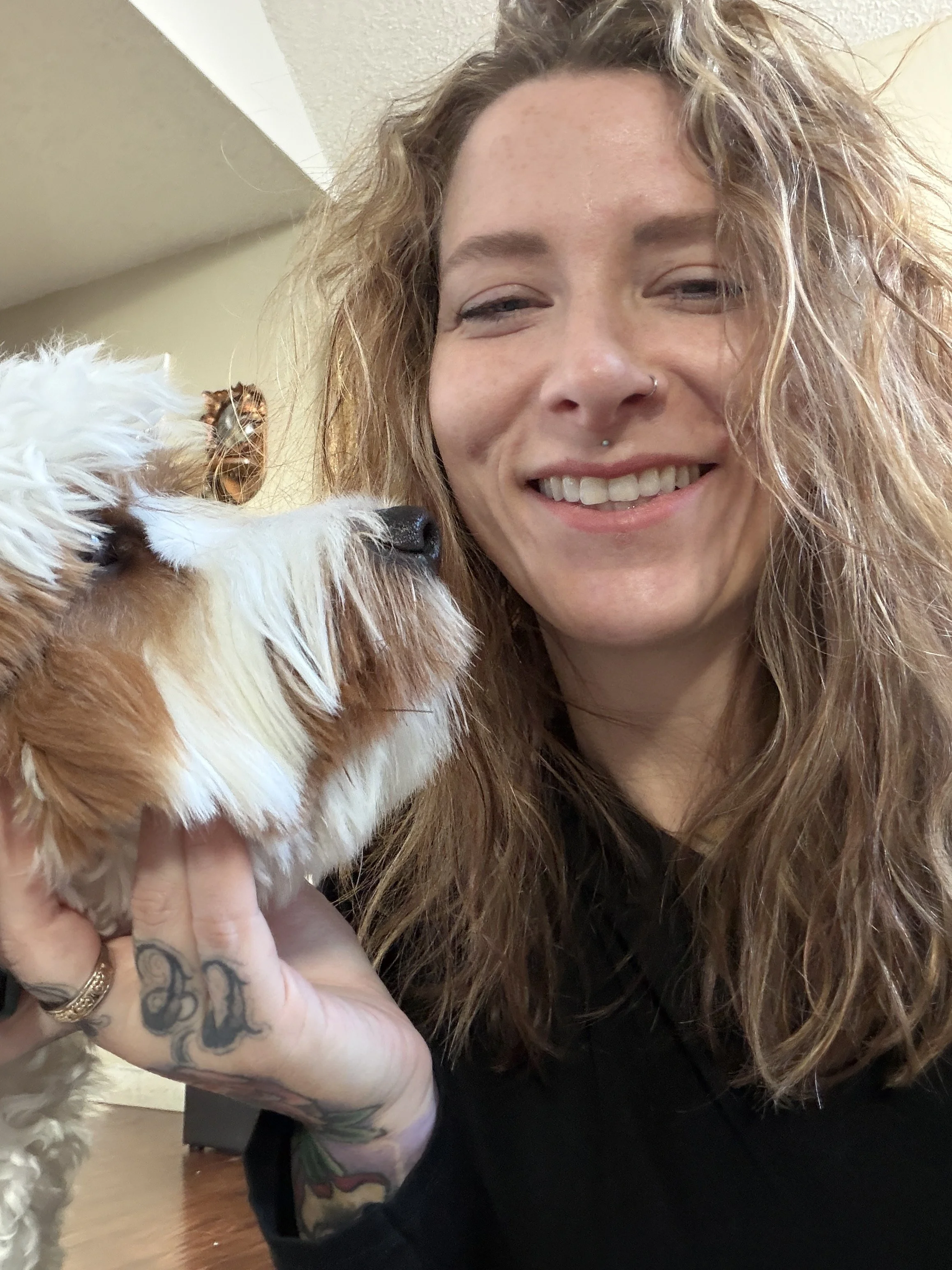 A woman with curly red hair smiling and holding a small, brown and white puppy with fluffy fur.