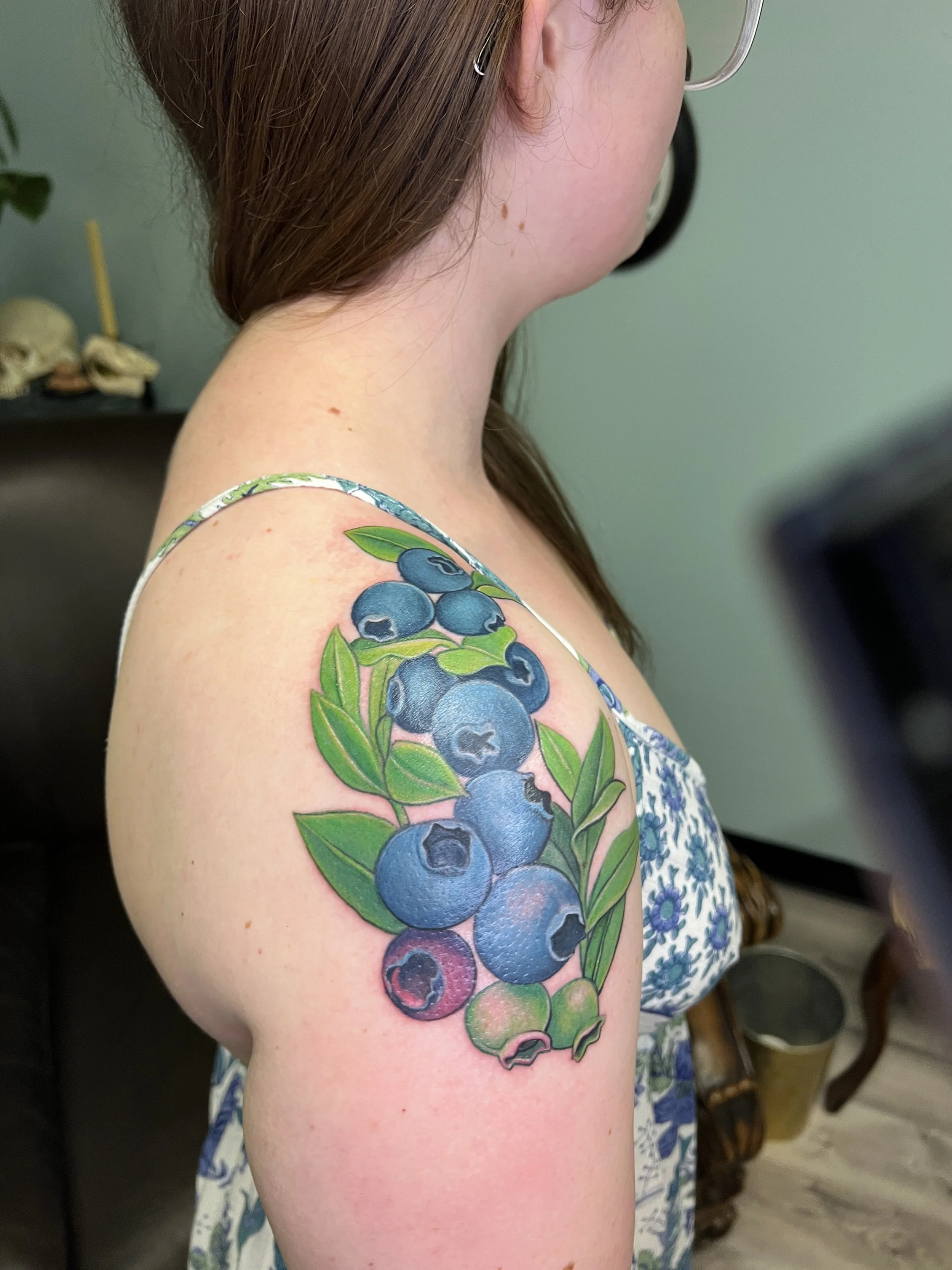A woman with fair skin and straight brown hair wearing glasses and a dress with a blueberry and green leaf tattoo on her upper arm.