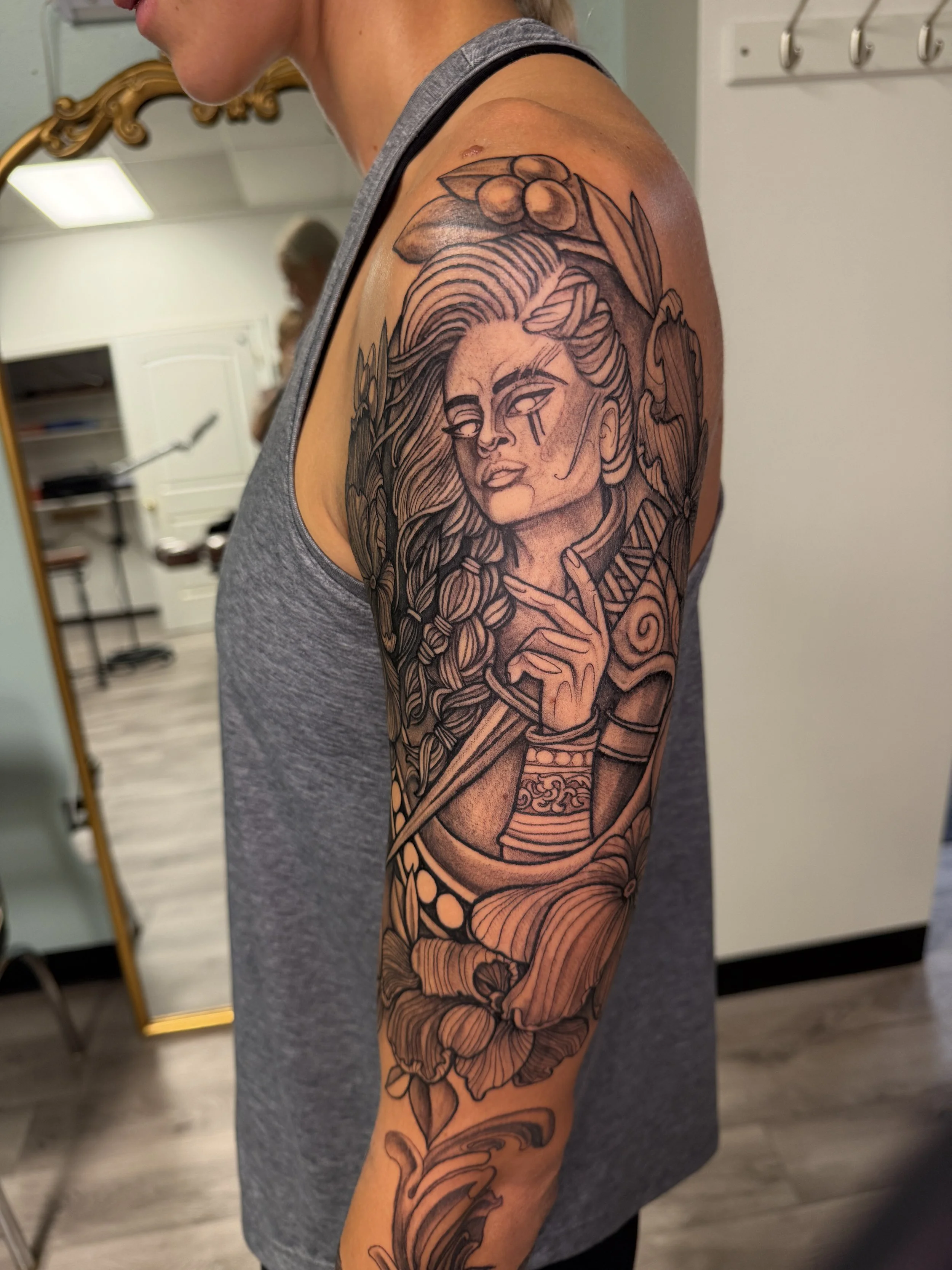 Tattoo of a woman with long hair, holding a bow, with berries and leaves above her head, on the person's upper arm.