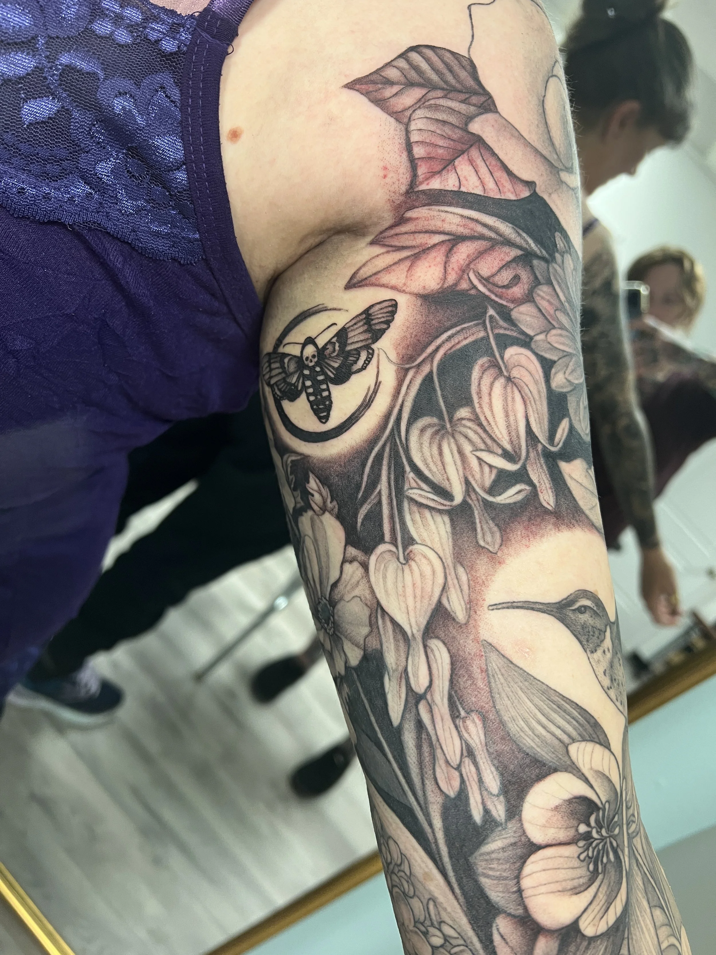 Close-up of a person's arm with a detailed black and gray floral and nature-themed tattoo, including leaves, flowers, a moth with a skull on its body, and a humming bird.