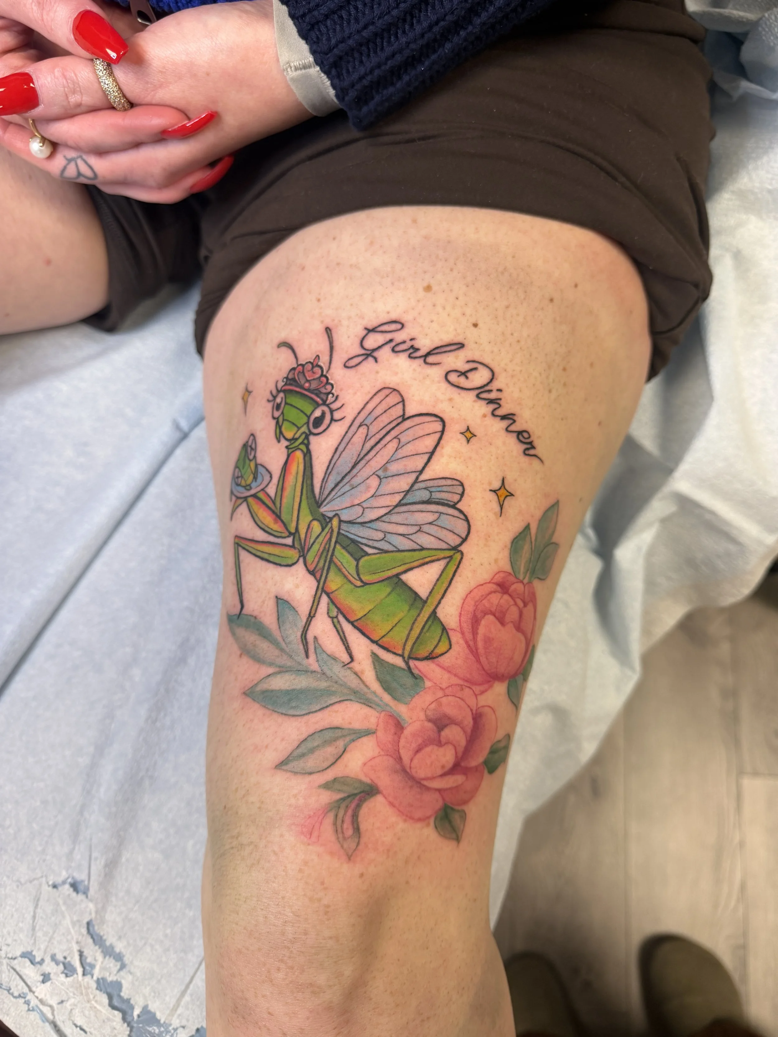 A colorful tattoo of a praying mantis with butterfly wings on a person's thigh, accompanied by the phrase 'Girl Power' in script and small stars, with pink flowers and green leaves underneath.