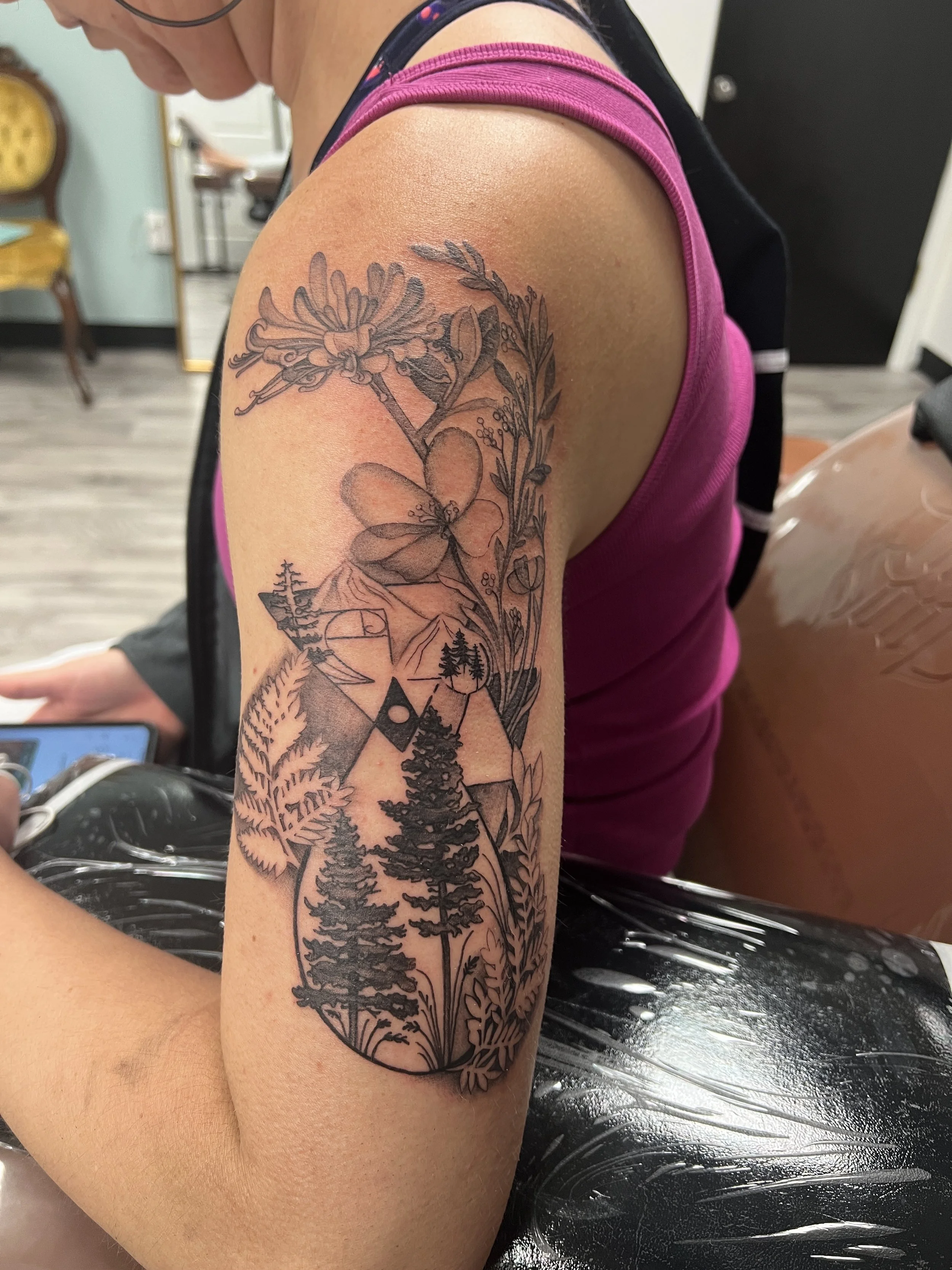 A person with a detailed black and gray tattoo sleeve on their upper arm featuring trees, mountains, a heart, and floral elements. 