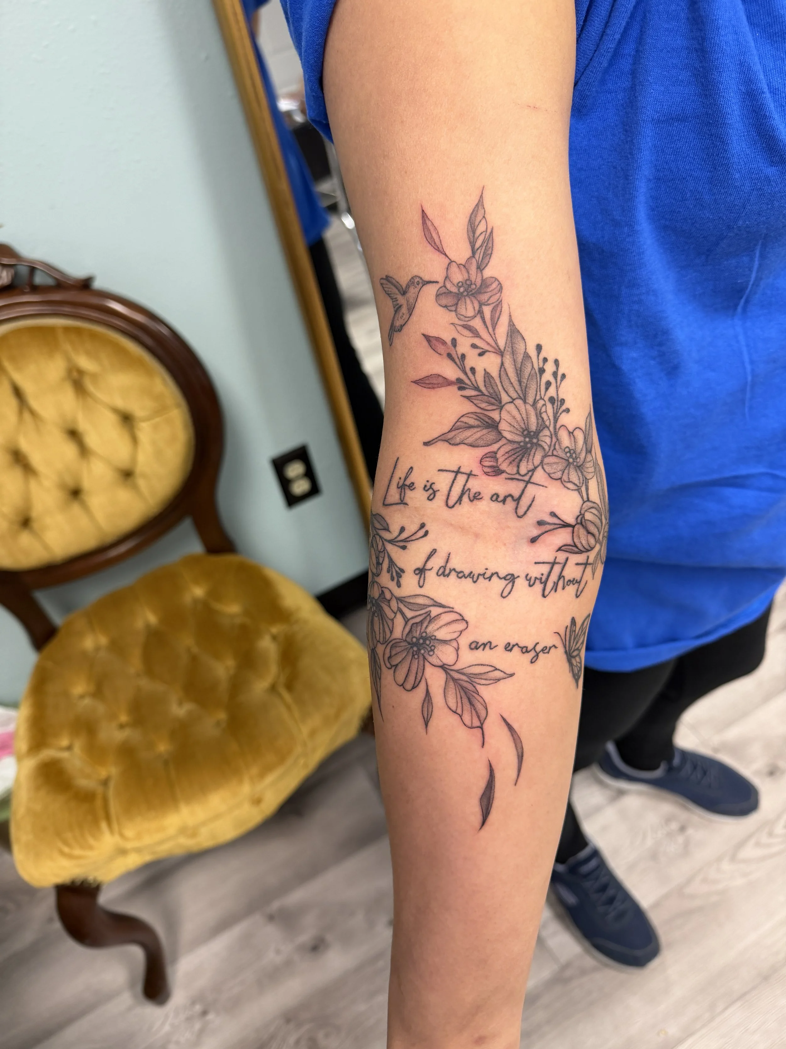 A person's arm with a black ink floral tattoo featuring large flowers, leaves, and a hummingbird, along with cursive text that reads "life is the art of drawing without an eraser." The background shows a vintage yellow upholstered chair and part of a