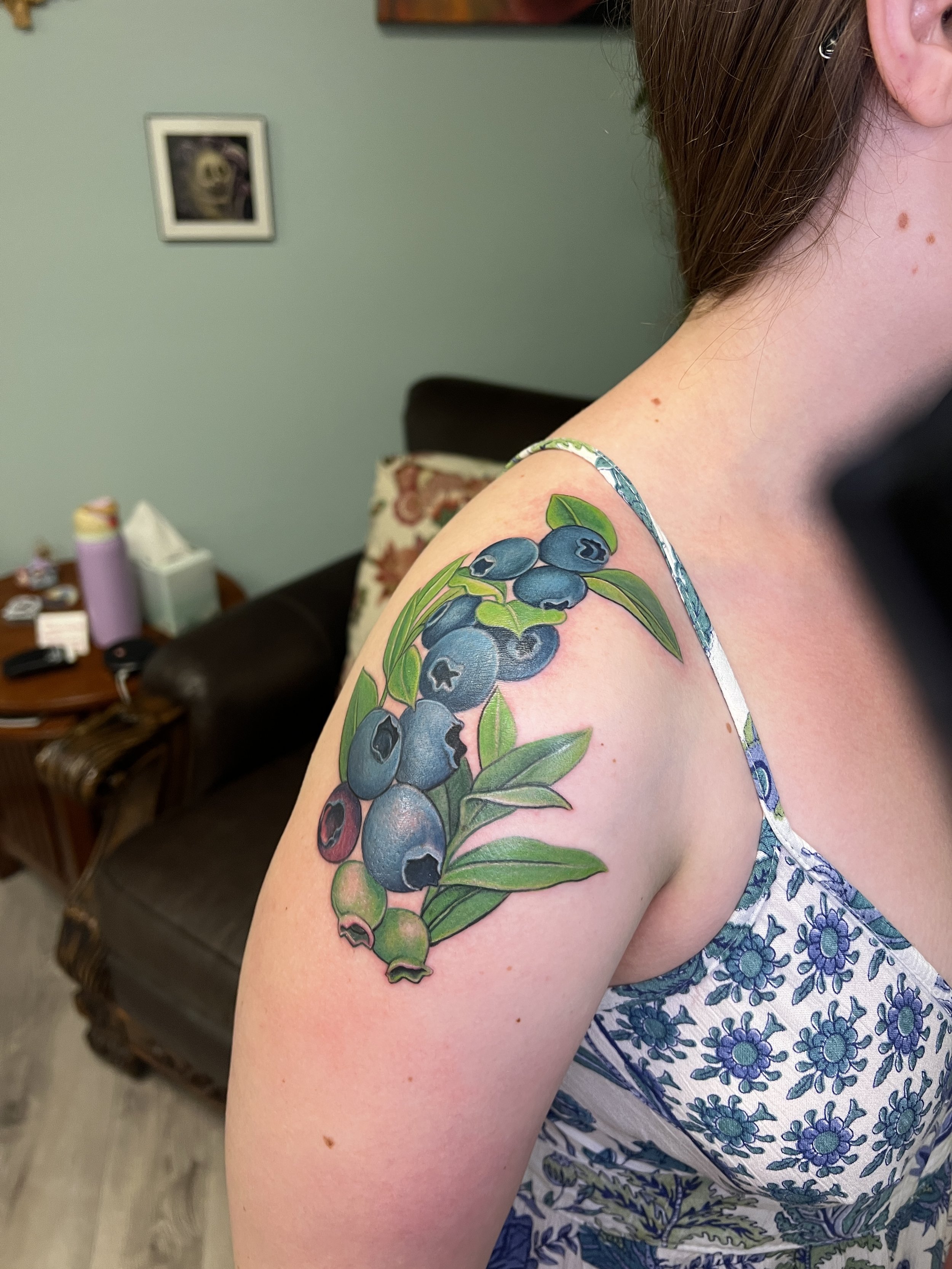 A woman's shoulder with a colorful tattoo of blueberries and leaves. She wears a sleeveless dress with a blue floral pattern. In the background, there's a living room with a brown sofa, a side table, and a picture on the light green wall.