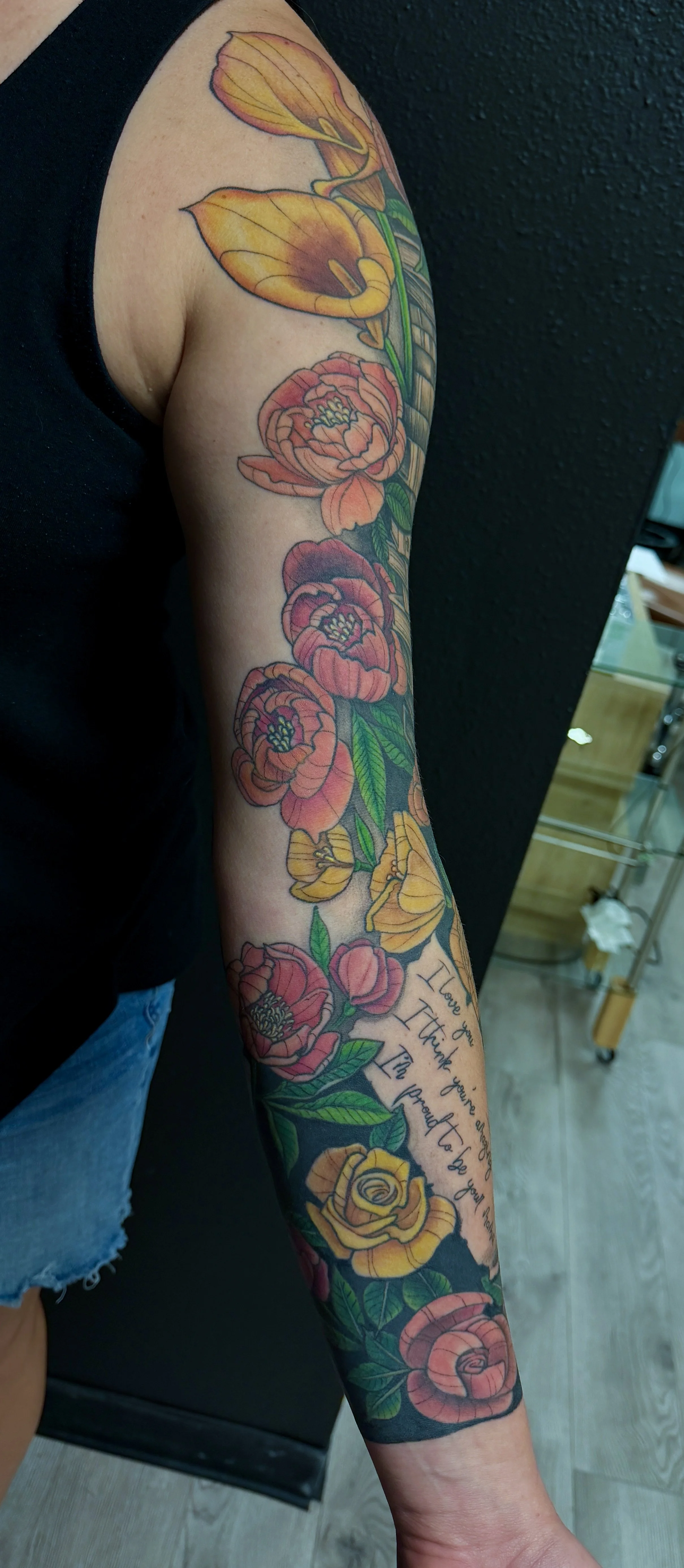 Colorful floral tattoo sleeve on person's arm, featuring large pink and yellow flowers with green leaves and a quote.