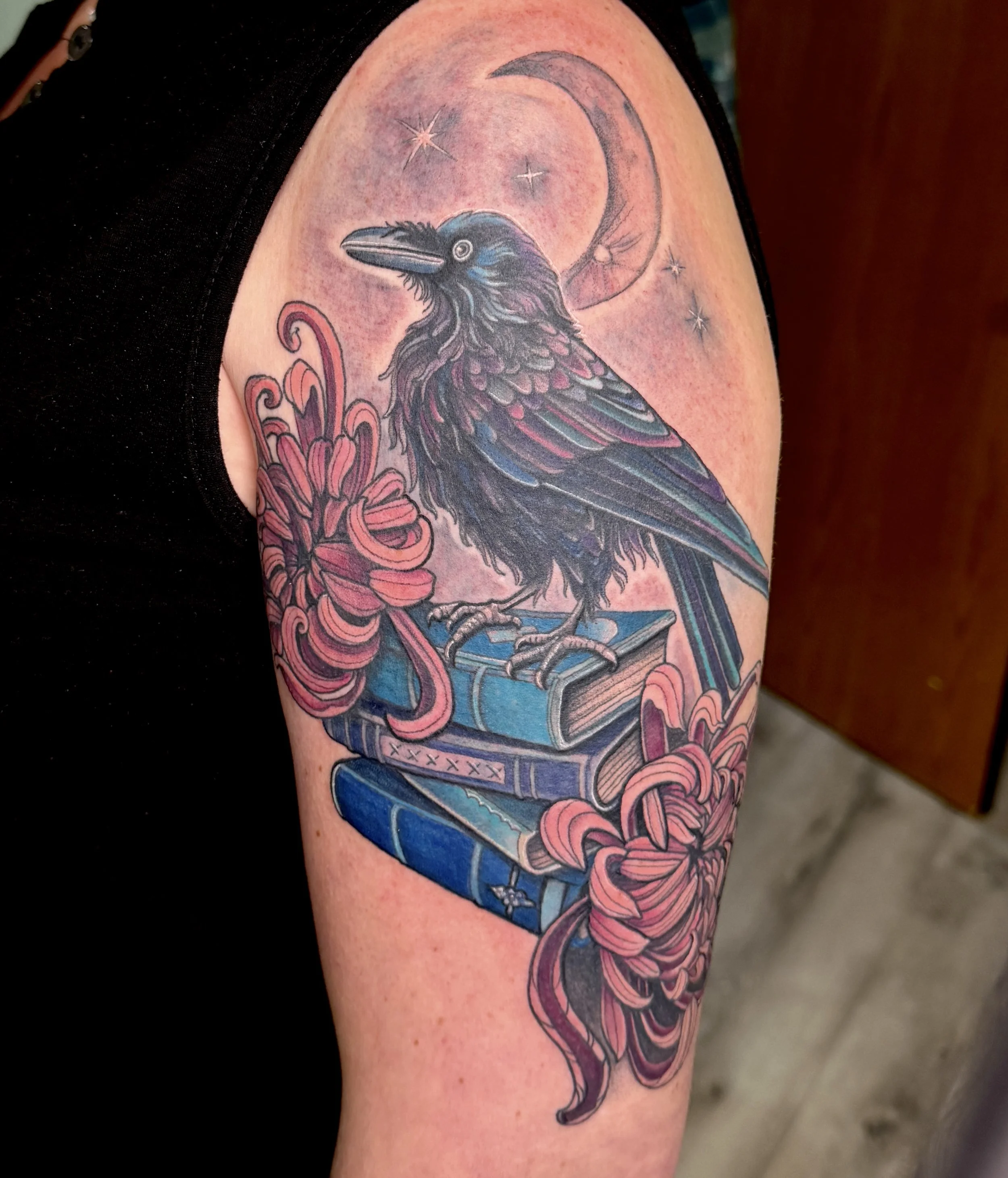 Tattoo of a raven with detailed feathers, a crescent moon, and sparkles in the background, with pink floral elements and blue sky.