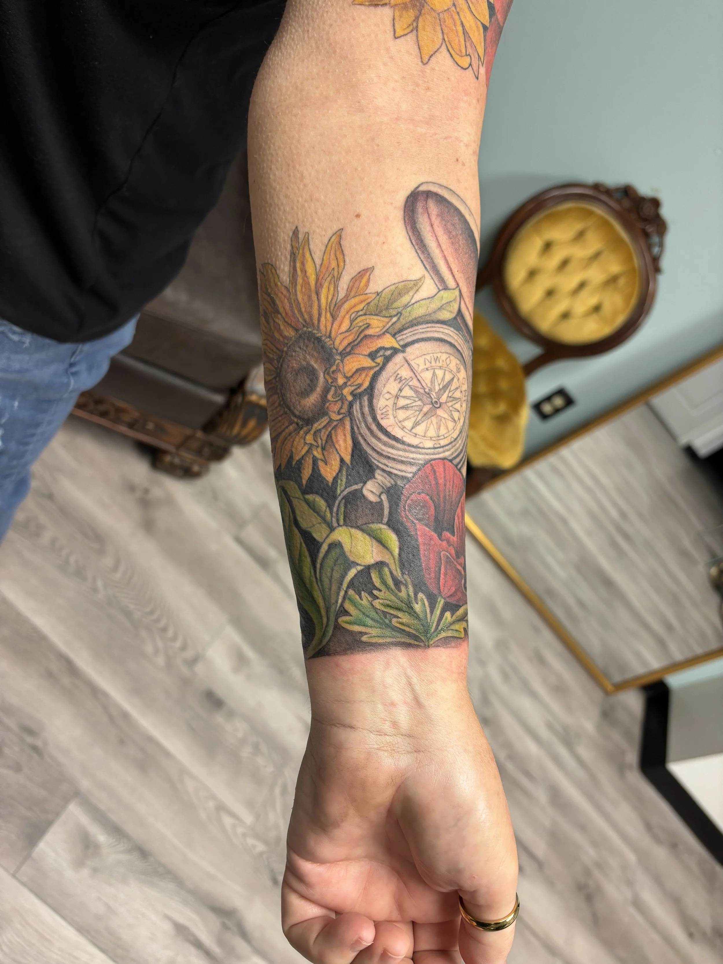 A person's arm with a colorful tattoo featuring a sunflower, a seashell, and other botanical elements.