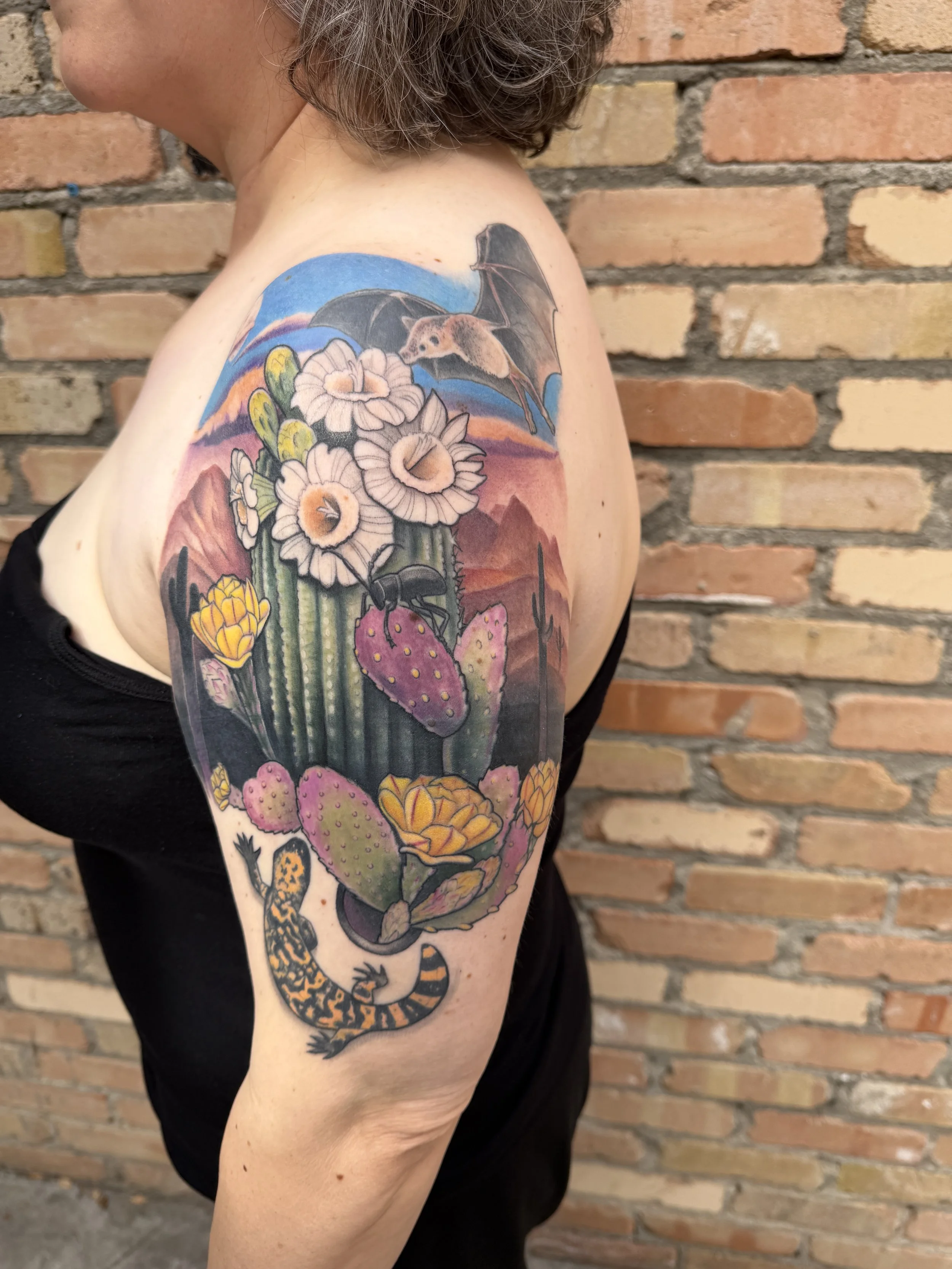 A person with a colorful tattoo sleeve of desert plants, a bat, and a jaguar standing against a brick wall.
