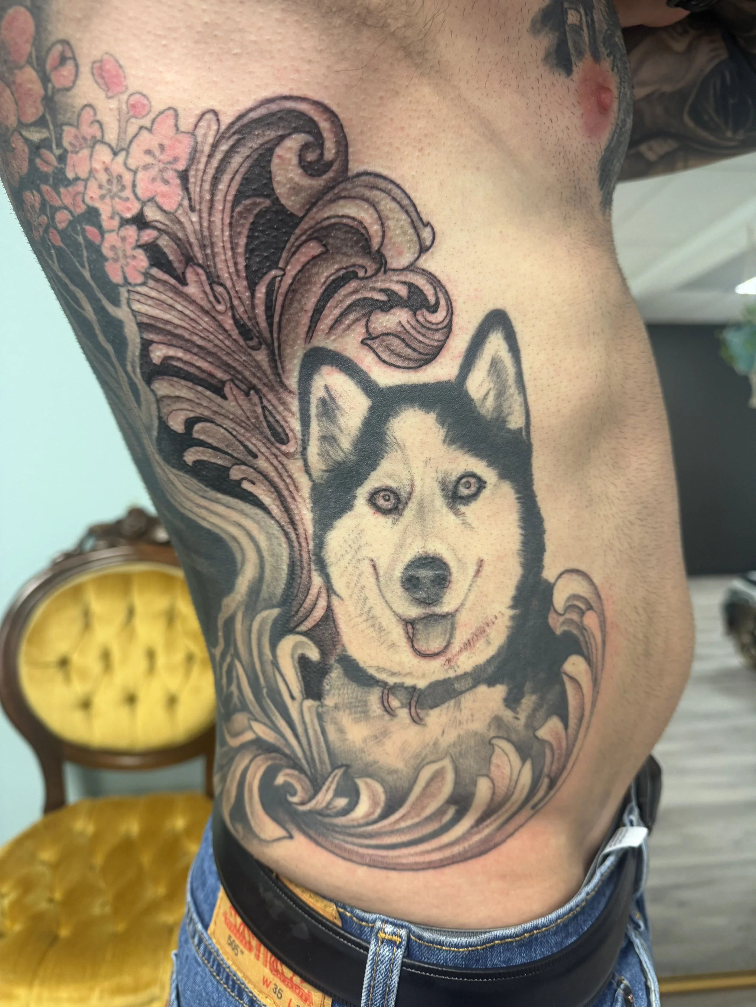 Tattoo of a husky dog's face on a person's side, with design elements resembling swirling waves and cherry blossoms nearby.