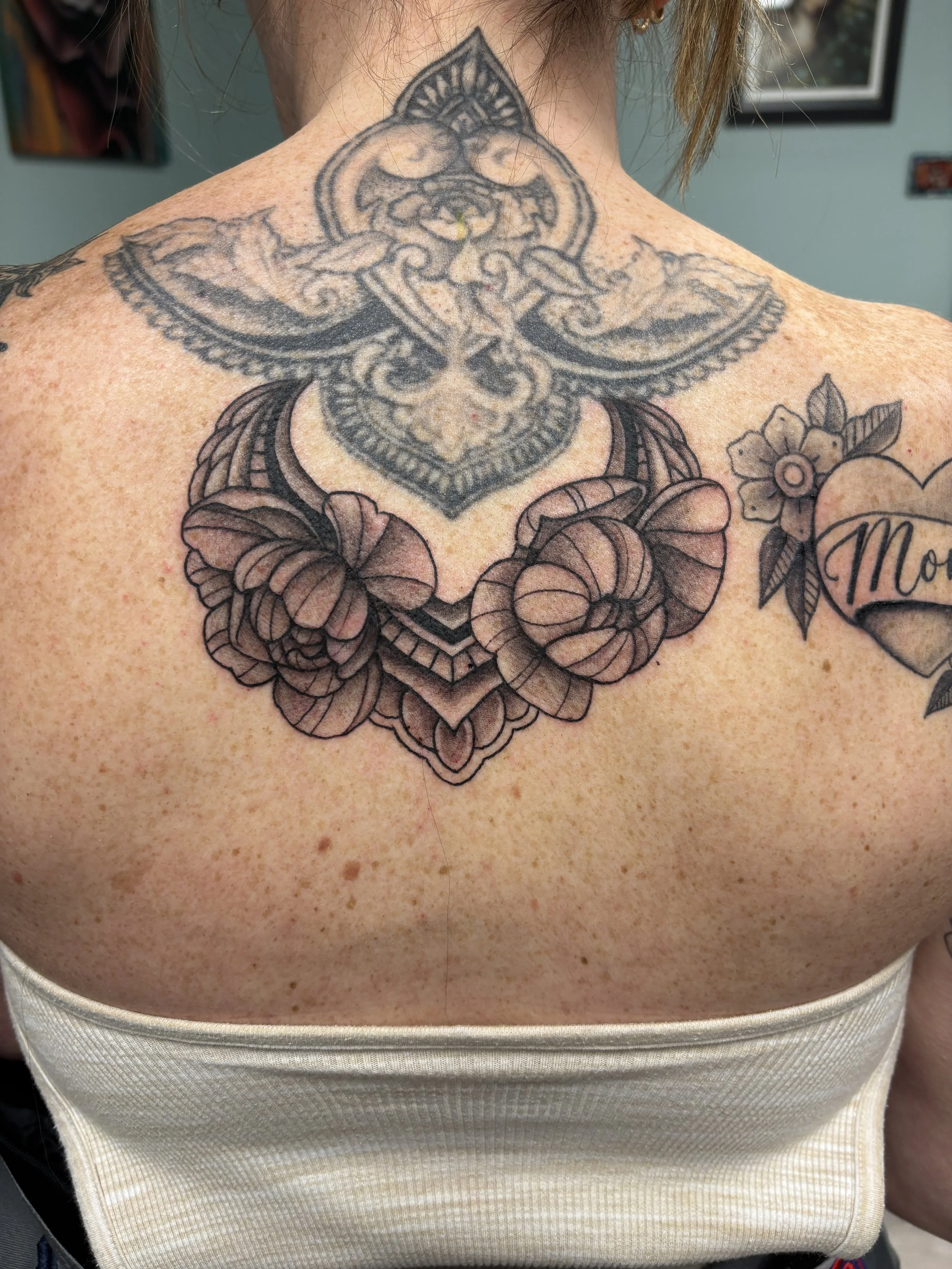 Close-up of a person's upper back and shoulders showcasing a large, intricate tattoo of a stylized owl with floral elements, primarily in black and gray ink.