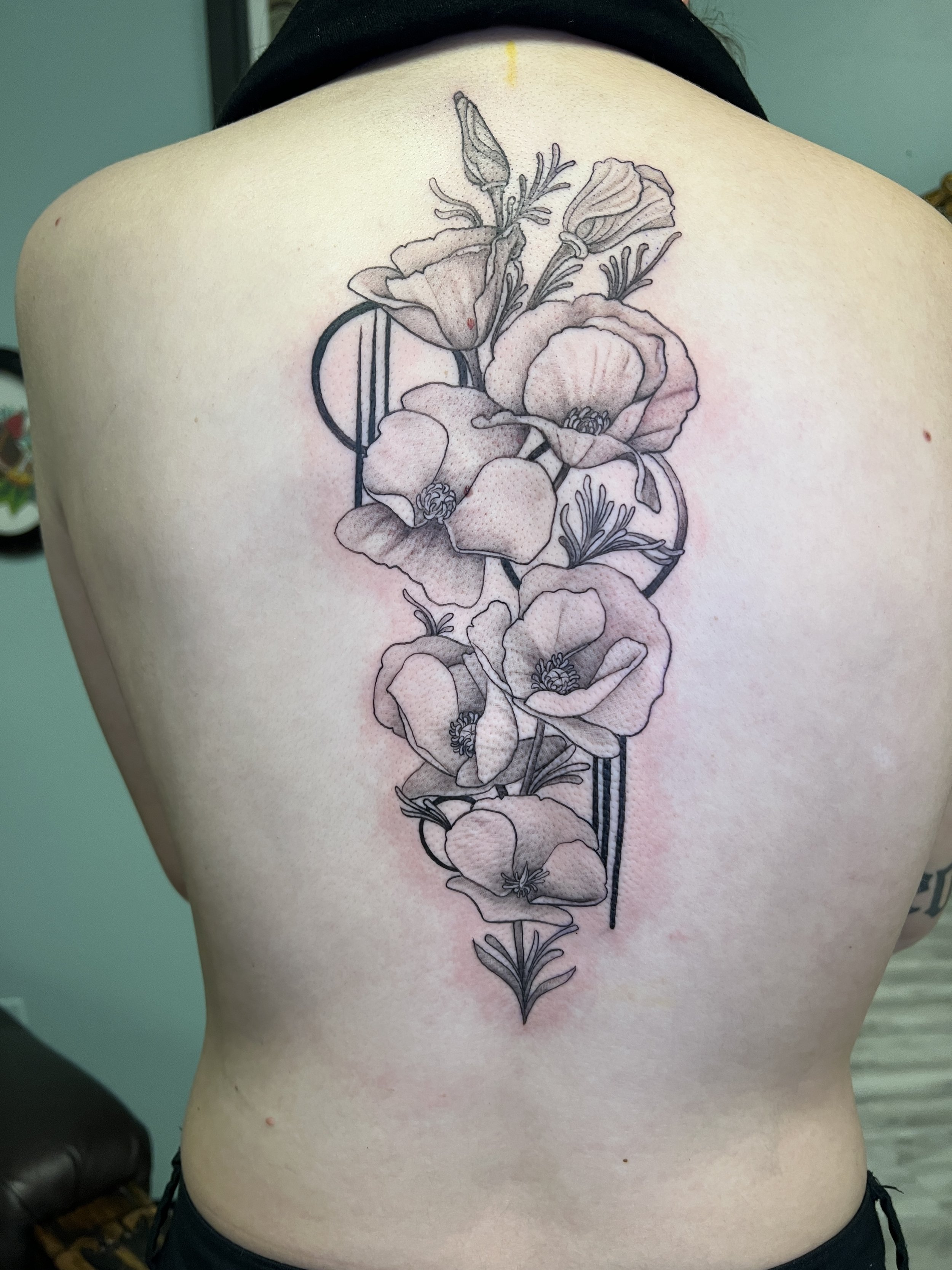 Black and white tattoo of a floral design on a person's upper back, featuring large open flowers with detailed petals and stamens, connected by a stylized circular element.