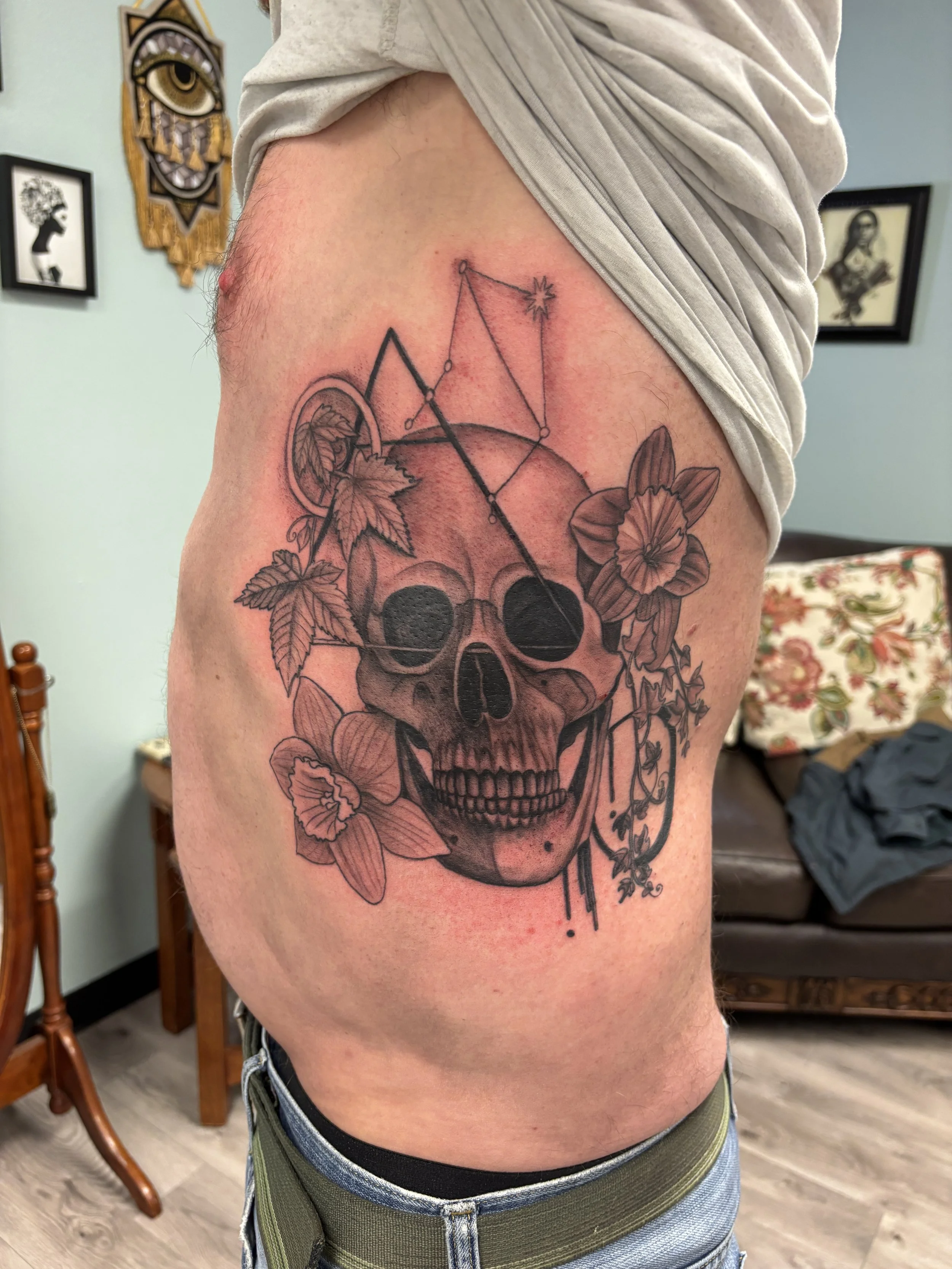Tattoo of a skull with flowers and leaves, with geometric designs and lines, on a person's side torso.