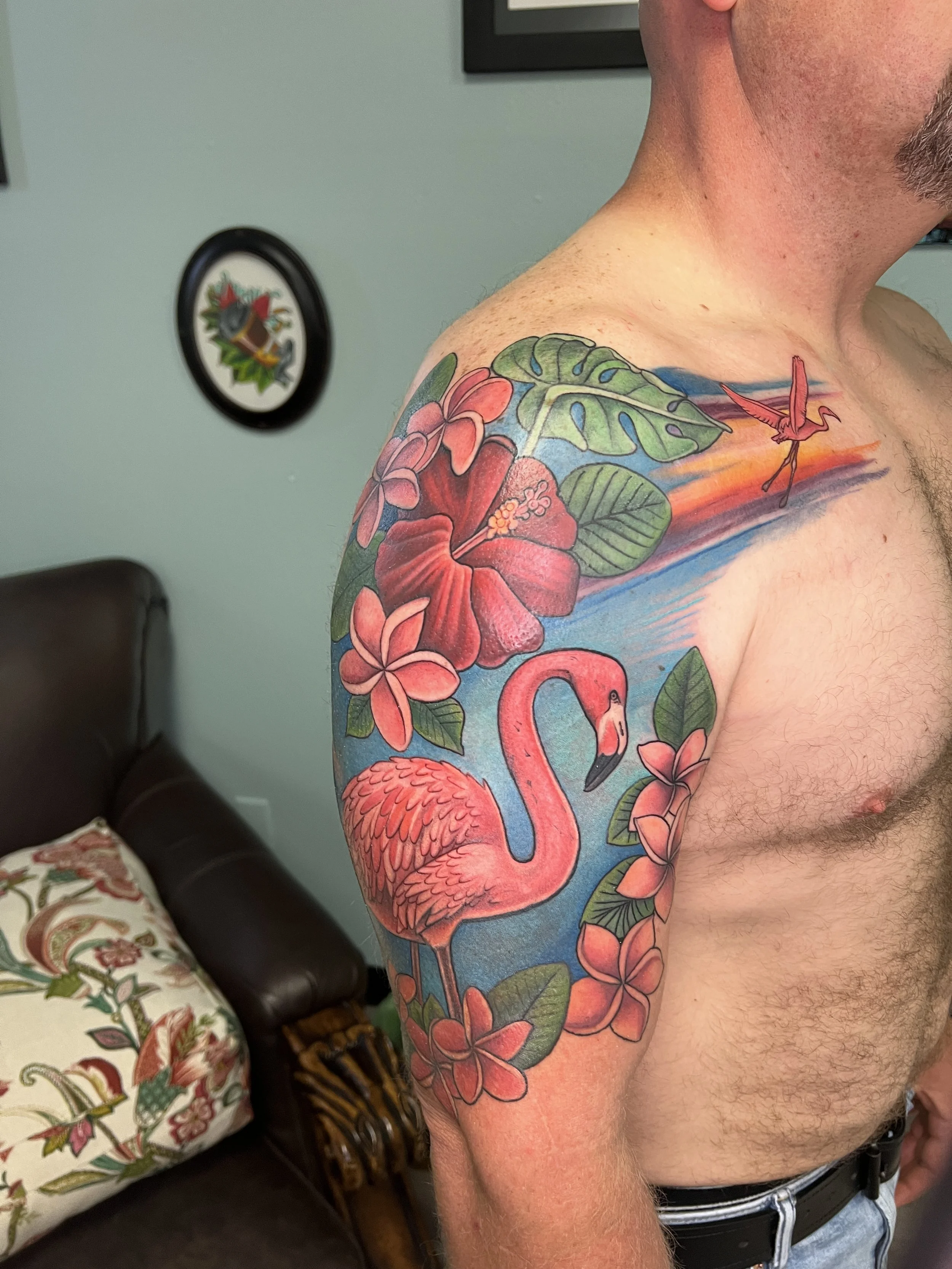 A person's upper arm and shoulder featuring a colorful tattoo of pink and red flowers, green leaves, and a flamingo.