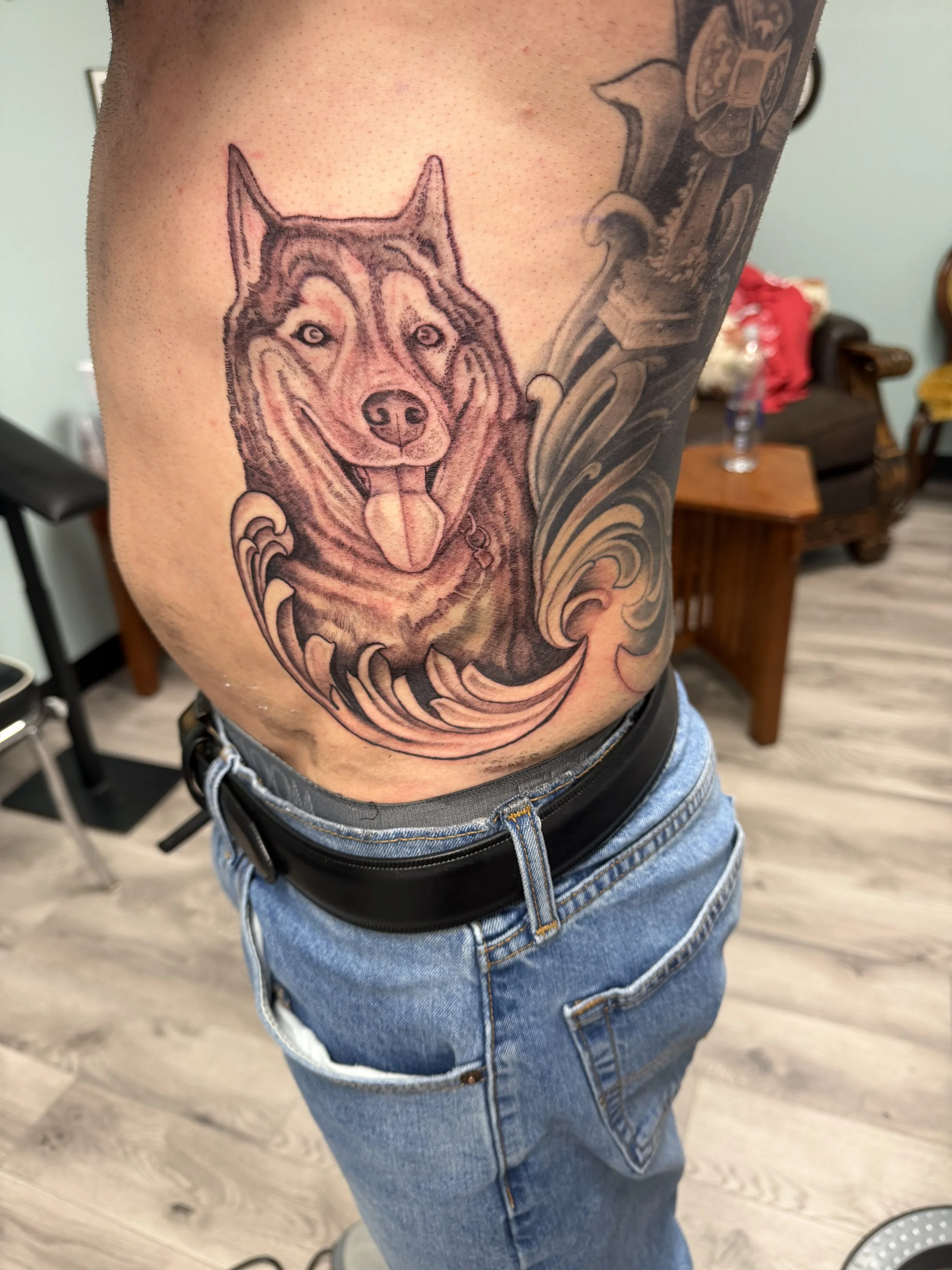 Tattoo of a dog's face with a swirling design on a person's side, wearing jeans and standing indoors.