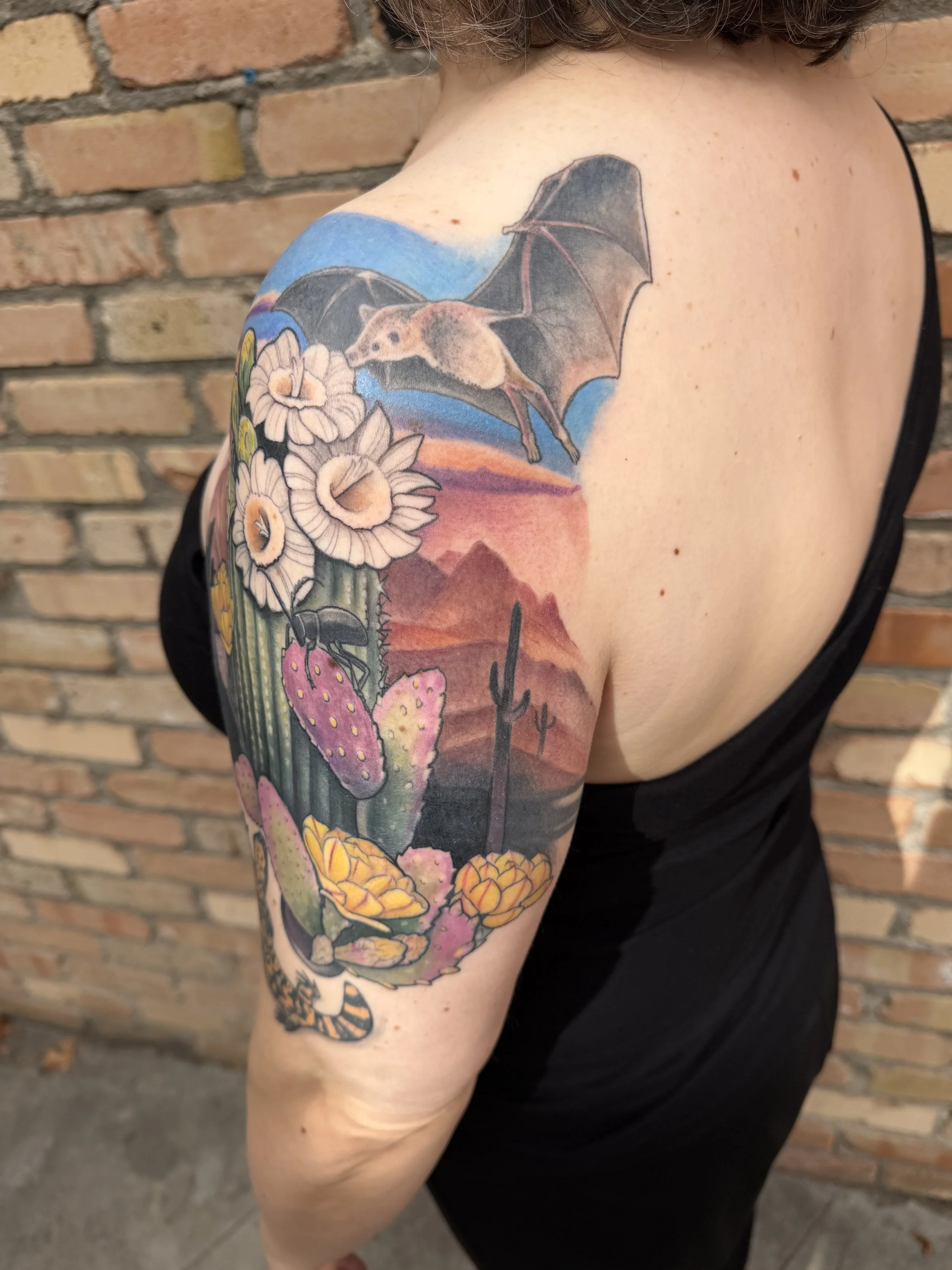 Colorful tattoo sleeve featuring a flying bat, desert landscape, cactus, and flowers on a person's upper arm against a brick wall background.